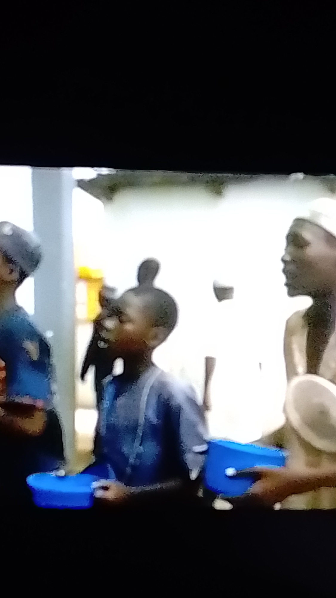 The image shows a group of children, likely in Nigeria. One child in the foreground, wearing a blue garment, is holding a blue object, possibly a container or toy, in their hands. Another child to their right is also holding a similar blue object. In the background, other children and adults are visible, some wearing white head coverings. The environment appears to be outdoors, possibly a street or courtyard, with buildings and greenery in the distance. The image is blurry and has a low resolution, making it difficult to discern fine details.