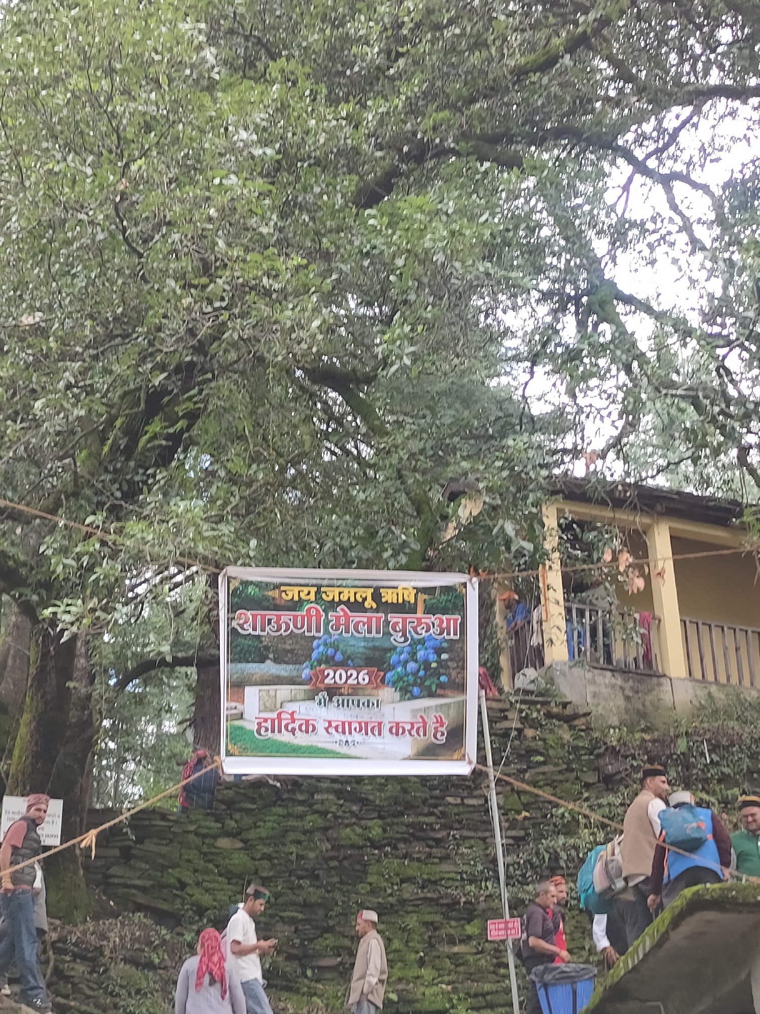 In Majhach, India, a banner hangs from a tree displaying "Jai Jamlu Rishi Shajuni Mela Burua 2026". The banner features an image of blue hydrangeas and reads, "We welcome you." Several people are visible in the scene. In the foreground, a man in a red scarf and a white shirt walks up stone steps. To his left, another man in a red scarf and grey shirt stands near a trash bin. Further up the steps, a man in a white shirt and blue jeans is looking at his phone, and a man in a light brown jacket and white shirt stands near him. To the right, a man in a brown vest and a blue backpack is walking up the steps. In the background, a building with yellow walls and a dark roof is visible, with a railing along its veranda. The environment is lush and green with a large tree in the foreground and stone walls lining the path.