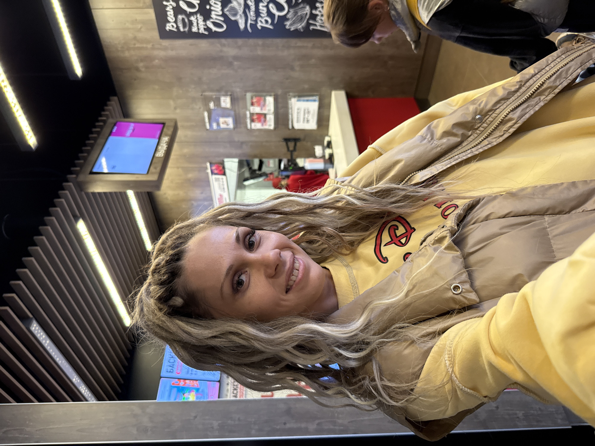 A woman with blonde dreadlocks, wearing a yellow sweatshirt with a red logo and a beige puffer vest, smiles as she takes a selfie. The background appears to be a fast-food restaurant, with a digital menu board displaying the word "ВЫДАЧА" (meaning "issue" or "collection" in Russian) and other menu items. A child with brown hair is visible in the lower right corner of the image, facing away from the camera. The restaurant has wooden paneling on the walls and ceiling, with fluorescent lighting. The overall scene suggests a casual dining or takeout environment in Korobovo, Russia.