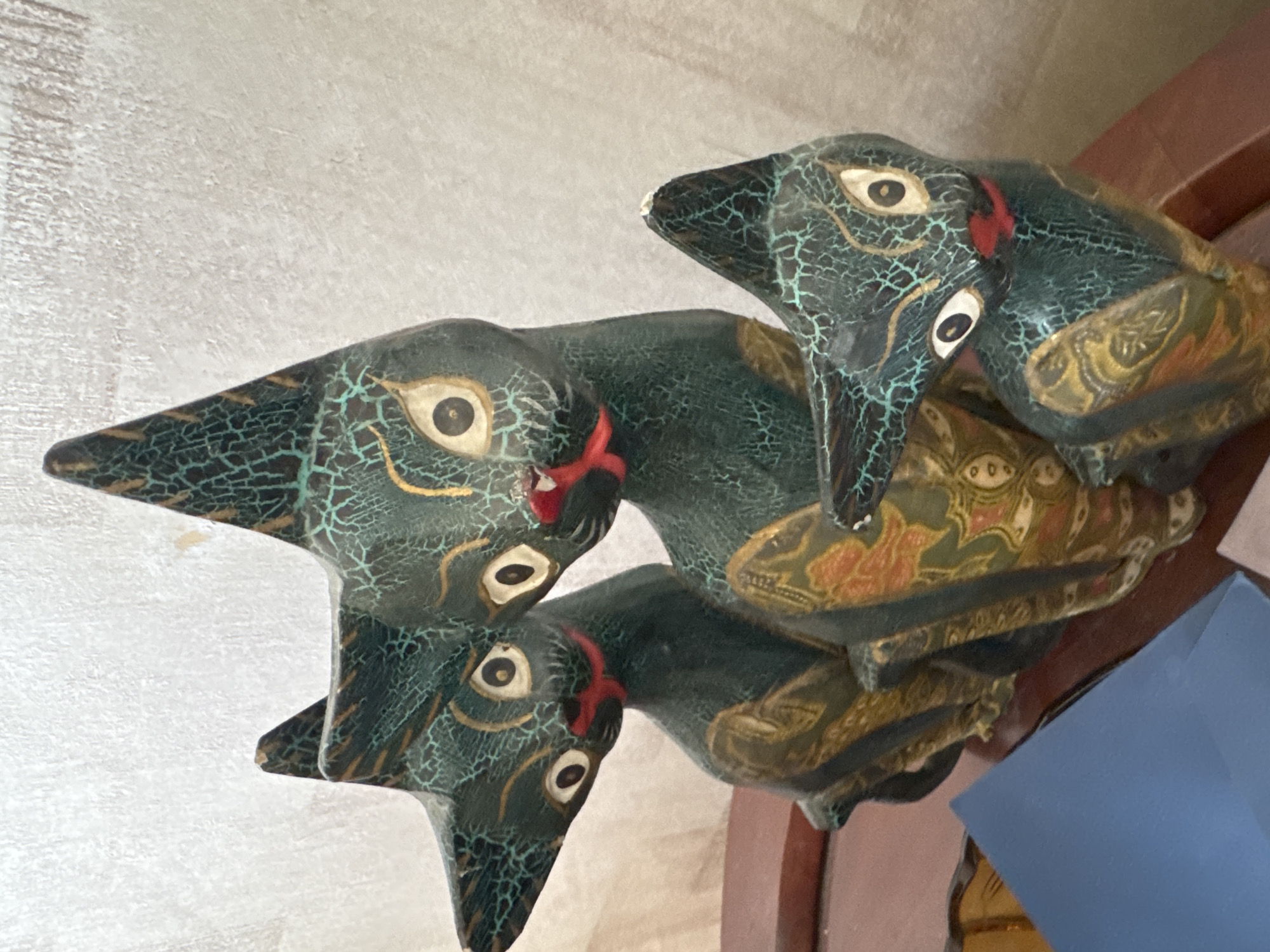 A close-up, slightly tilted photograph depicts a stack of three stylized, dark-colored wooden bird figurines. The figurines feature elongated necks and pointed beaks with a teal-cracked paint texture covering a darker base. The eyes are large and stylized, and red paint highlights the area around the neck of each bird. The birds appear to be stacked on top of each other, with the top bird facing slightly to the right. The figures are patterned with orange and gold details. The background consists of a light-colored textured wall on the left and a portion of a wooden surface on the right, with a blue rectangular object partially visible. There is no evidence that these are related to Sofia, Bulgaria.