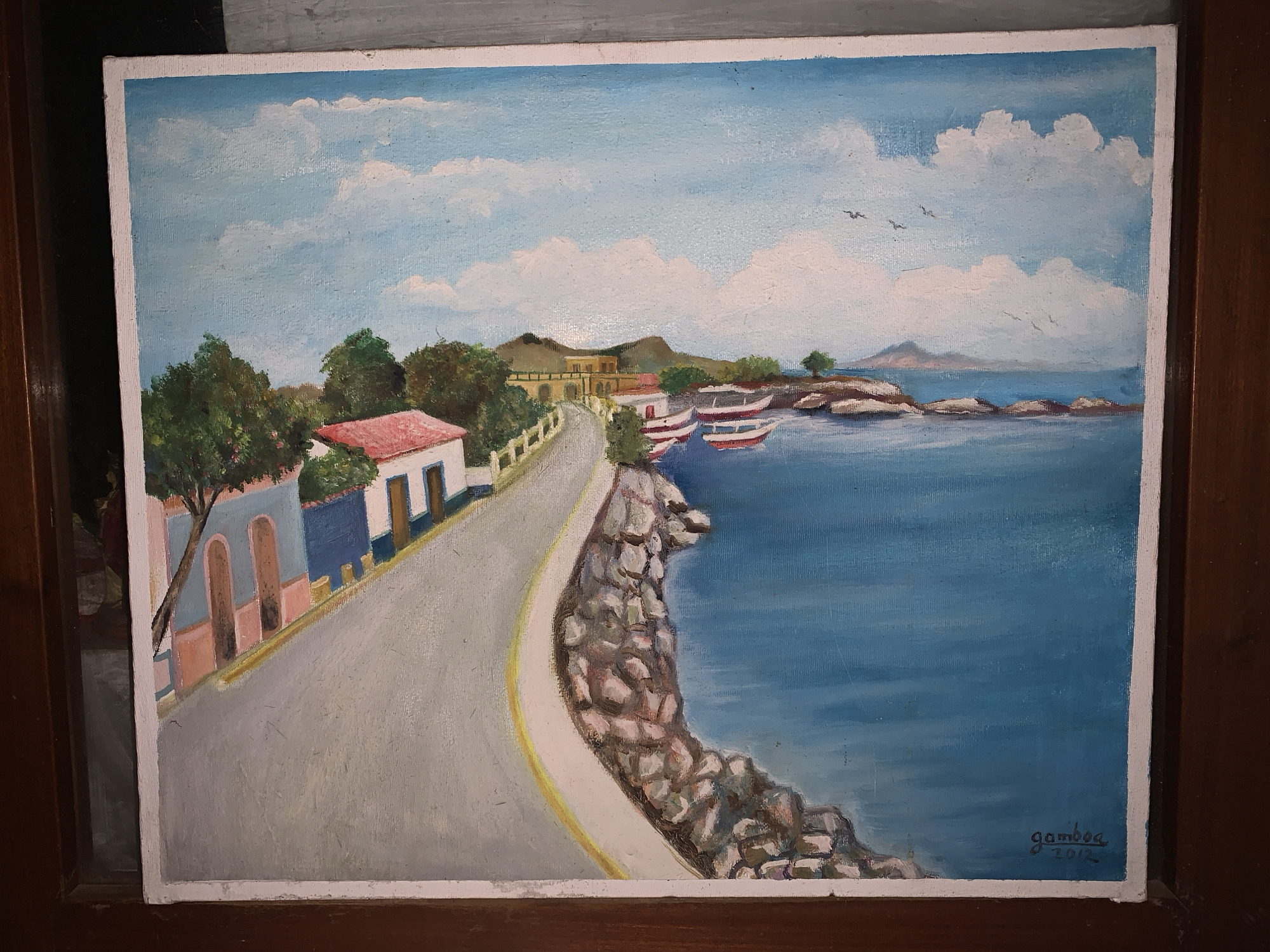 The image shows an oil painting depicting a coastal scene with a road winding along the shoreline. On the left side of the road are buildings with a variety of colors, including blue, white, pink and red. A rocky cliff separates the road from the ocean. In the water, there are a few boats, and in the background, there is land with houses and mountains. The sky is blue with some scattered white clouds and birds flying. The signature "gamboa 2012" is visible in the lower right corner. The scene appears to represent a coastal community with a road running alongside the waterfront. The location could be Santa Ana, Venezuela, as implied, considering its geography.