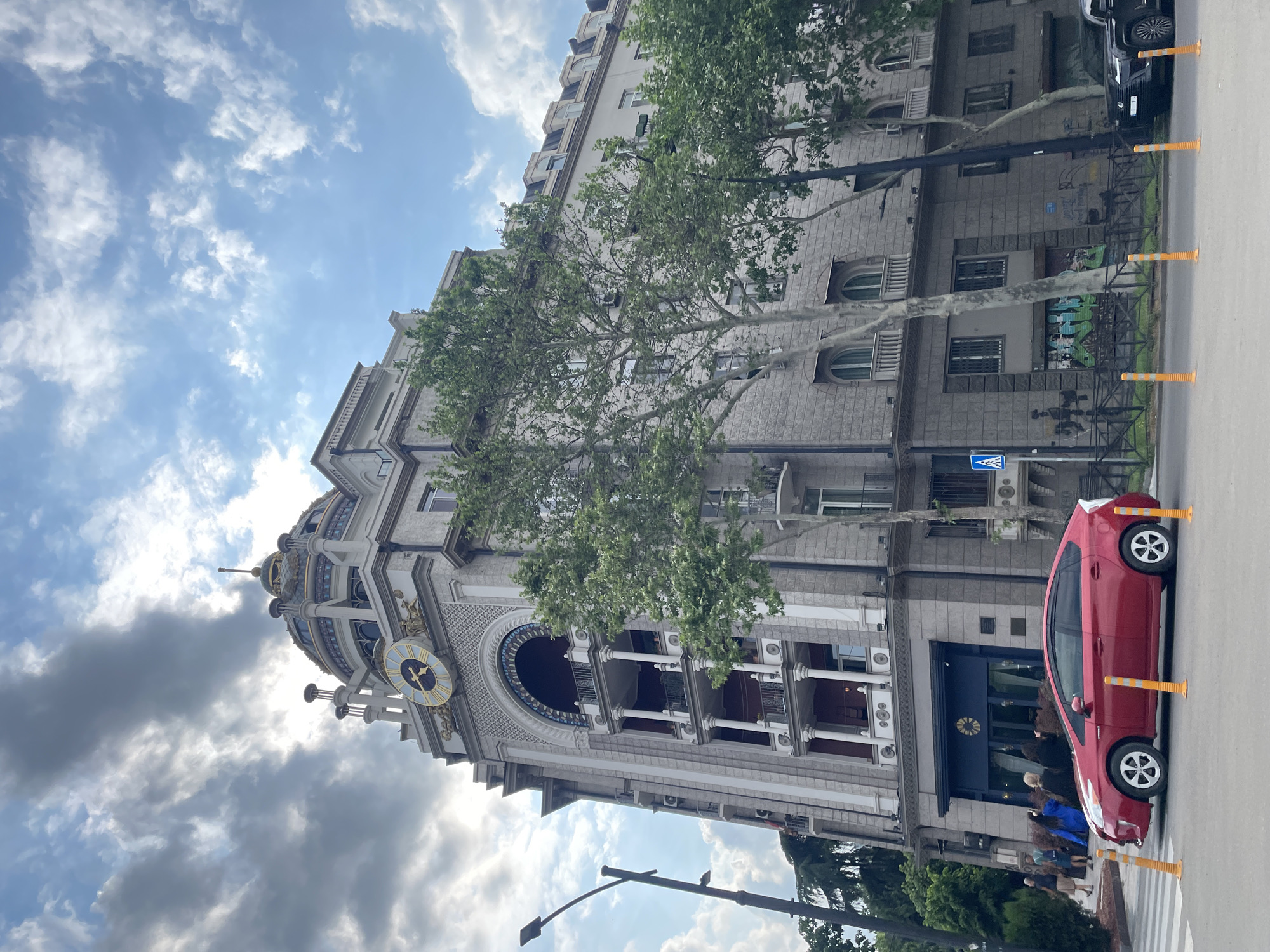 The image shows a street view in Tbilisi, Georgia, featuring a distinctive building with a clock tower. The architectural style suggests a historical or landmark structure. A red car is parked along the sidewalk, close to the building's entrance, which appears to be a restaurant or business. People are seen near the entrance. The sky is partly cloudy, with sunlight illuminating the scene. Orange traffic cones are present along the curb.