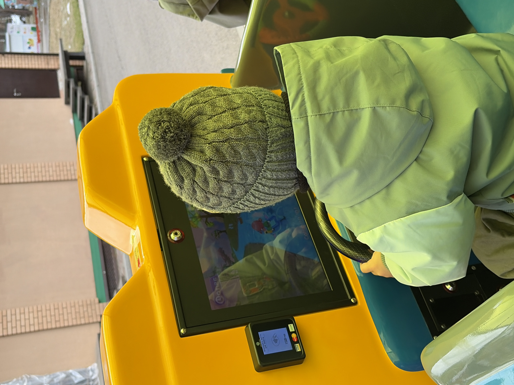 A person in a gray knit hat and light green jacket is seated in a yellow and blue ride-on machine. The machine has a black-framed screen displaying a cartoon animation and a black payment terminal with an illuminated screen. The scene is outdoors, possibly in an amusement area, as suggested by the ride and the hint of surrounding structures. The exact location is Chaikovsky, Russia, but further environmental details are limited in the frame.
