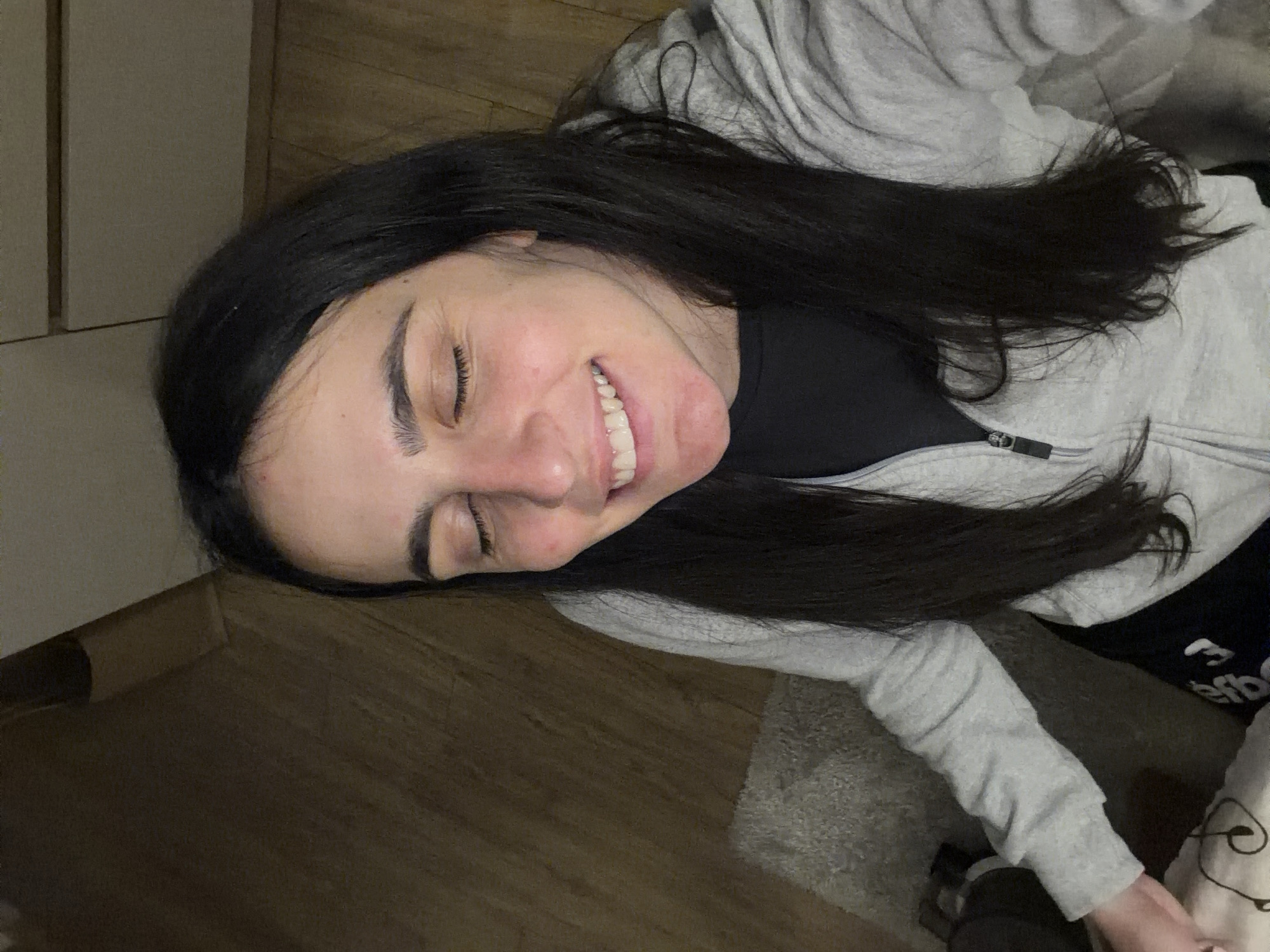A woman lies on a wooden floor, smiling with her eyes closed. She wears a grey hoodie over a black shirt. Her dark hair is spread around her head. The background is blurry and indistinct, but suggests a simple interior setting. There is no visible information to determine a specific location in Sofia, Bulgaria.