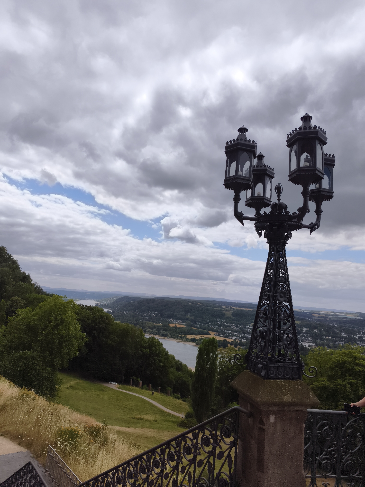 This image, taken in Königswinter, Germany, captures a scenic view from a high vantage point. In the foreground, a decorative, wrought-iron lamppost with multiple lanterns stands prominently. Its intricate design and dark metal contrast with the surrounding landscape. Below the lamppost, a stone pedestal supports its base, and a section of a decorative metal railing is visible.
The middle ground features a lush, green hillside with a pathway winding through it. Mature trees with dense foliage line the edge of the hill, and dry, tall grass grows in patches. In the distance, a wide river, likely the Rhine, flows through a valley. The river is bordered by hills covered in trees and dotted with buildings, suggesting a town or village.
The background is dominated by a dramatic sky filled with heavy, gray clouds, interspersed with patches of bright blue sky. The overall atmosphere is overcast, but with hints of sunlight breaking through. A hand holding a smartphone is visible on the right side of the image, suggesting someone is taking or has just taken this photograph. The image is shot from an elevated position, offering a panoramic perspective of the landscape.