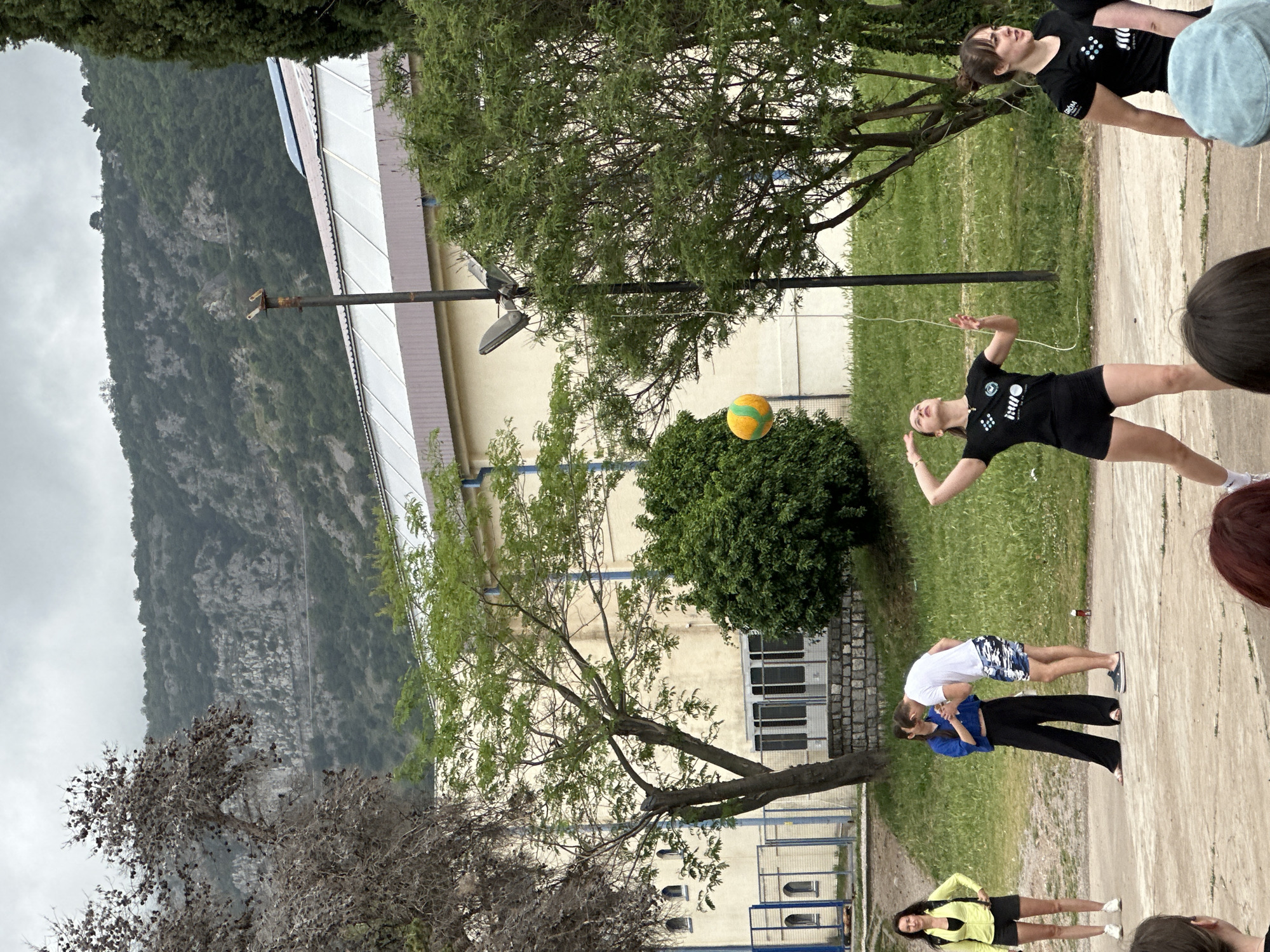 The image depicts a group of people participating in an outdoor activity on a paved area, likely a courtyard or recreational space in Budva, Montenegro.
A young woman wearing a black t-shirt and black shorts is in mid-action, reaching to hit a yellow and green ball. Behind her is a green bush and in the background is a building. To the left of the building is a mountain. To the right of the girl and ball are other people, including a girl in a black shirt and a man and woman doing a stretch.
The backdrop includes buildings, trees, a mountain, and a cloudy sky. The weather appears to be overcast. The scene is brightly lit.
