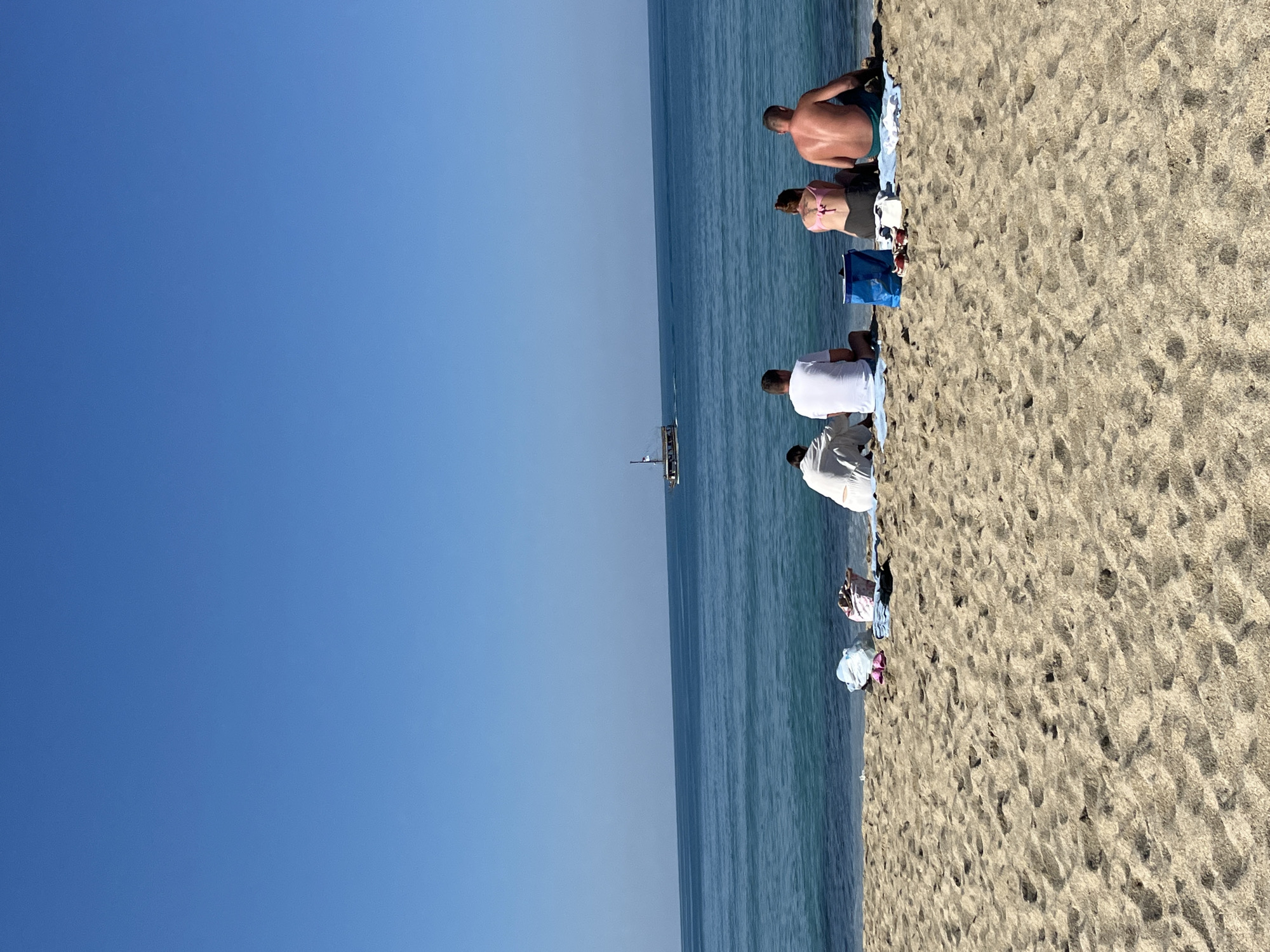 The image shows a beach in Alanya, Turkey. People are sitting on the sand near the water. There is a boat in the distance. The sky is clear blue.