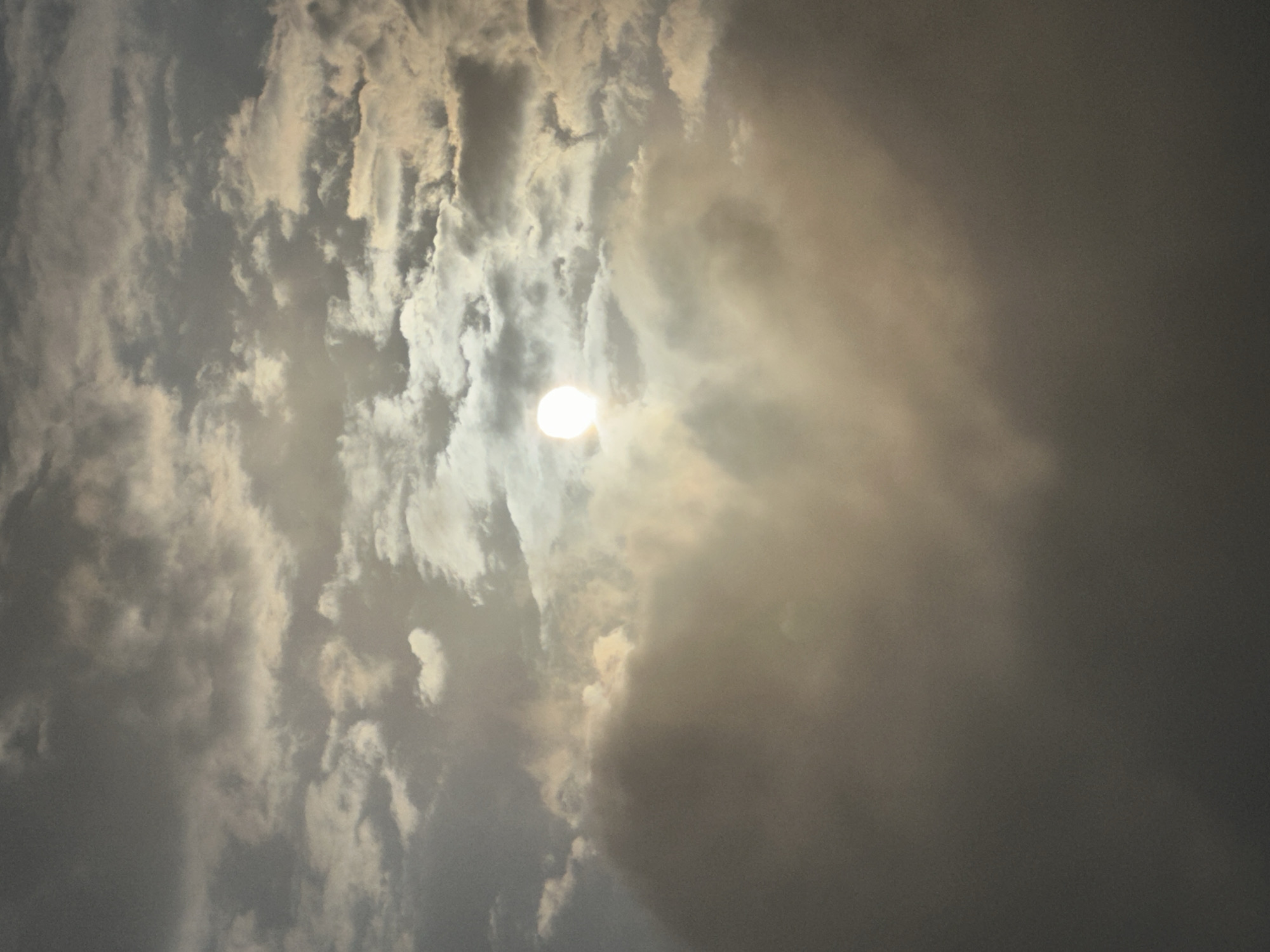The image depicts a sky filled with clouds, with the sun partially visible and diffused by the atmospheric conditions. The lighting suggests it might be dawn, dusk, or a heavily overcast day. The clouds vary in density and color, ranging from bright white where the sun's rays penetrate to darker grays and browns, indicating depth and varying thickness. There are no visible people, structures, or distinct geographical features that would specifically place the image in Estosadok, Russia, or any other city. The scene is exclusively focused on the sky and cloud formations.