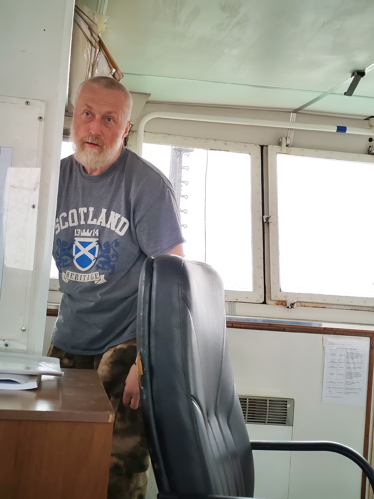 A middle-aged man with a white beard and short hair is pictured standing inside what appears to be the bridge or control room of a vessel. He wears a gray short-sleeved t-shirt with "SCOTLAND 1314 HERITAGE" printed on the front, featuring a Scottish Saltire flag and heraldic lion designs. His gaze is directed towards the camera with a neutral to slightly questioning expression.
The man is positioned behind a wooden desk, which holds some papers. A dark-colored office chair is prominently featured in the foreground to the right, partially obscuring his lower body and the camouflage-patterned trousers he is wearing. The background consists of large, multi-paned windows that provide bright, diffused daylight, suggesting it is daytime, possibly overcast.
The interior space is utilitarian, characterized by light-colored walls and ceiling. On the right-hand wall, behind the chair, an air vent is visible, and above it, a printed document is affixed. While the text on this document is not entirely legible, its format suggests it is an informational or procedural sheet, and given the location context of Shcheleiki, Russia, it is likely in Russian. The overall scene depicts a working environment on a maritime vessel.