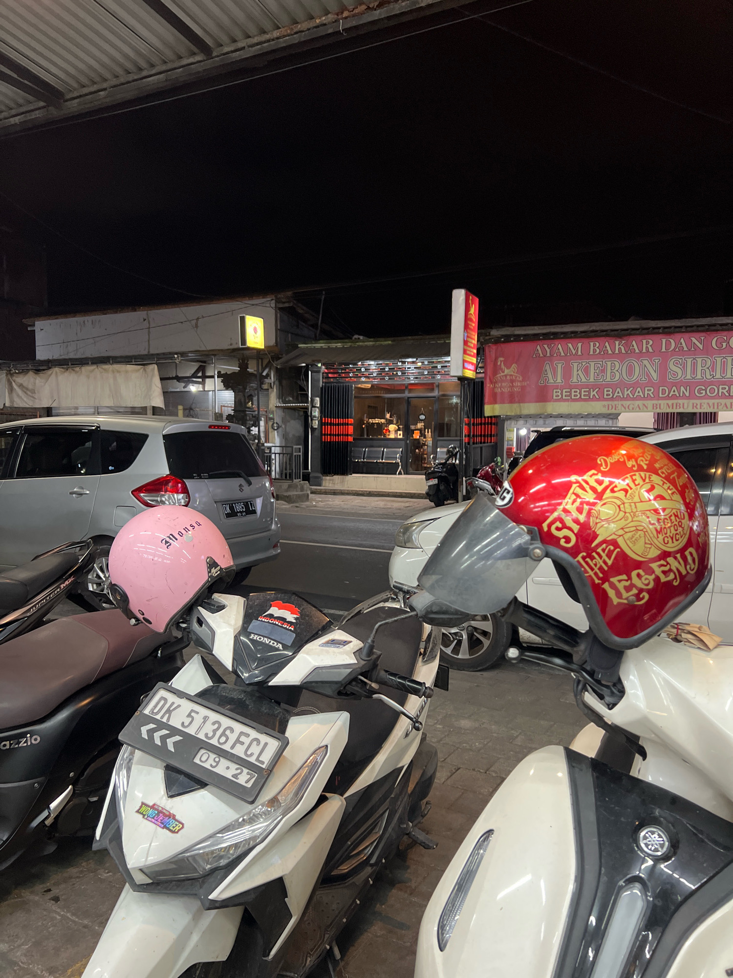 A street-level view at night shows several parked motorcycles in front of a building. On the left, a silver car is parked, and in the foreground, a black and white motorcycle is visible with a license plate that reads "DK 5136FCL" and "09 27". A white and black motorcycle is parked in front of it.
On the right, a red helmet with gold lettering, reading "STEVE THE LEGEND MOTOR CYCLE", is placed on the handlebars of a white motorcycle. Another motorcycle, black and brown, is parked to the left of the silver car.
A banner with red and yellow lettering, which appears to advertise "AYAM BAKAR DAN GORENG AI KEBON SIRIH", is visible above the building's entrance. The building itself has a modern storefront with glass doors and windows, revealing seating inside. The scene is illuminated by artificial lights from the building and streetlights. The overall impression is a busy street or parking area outside a food establishment in Denpasar, Indonesia.