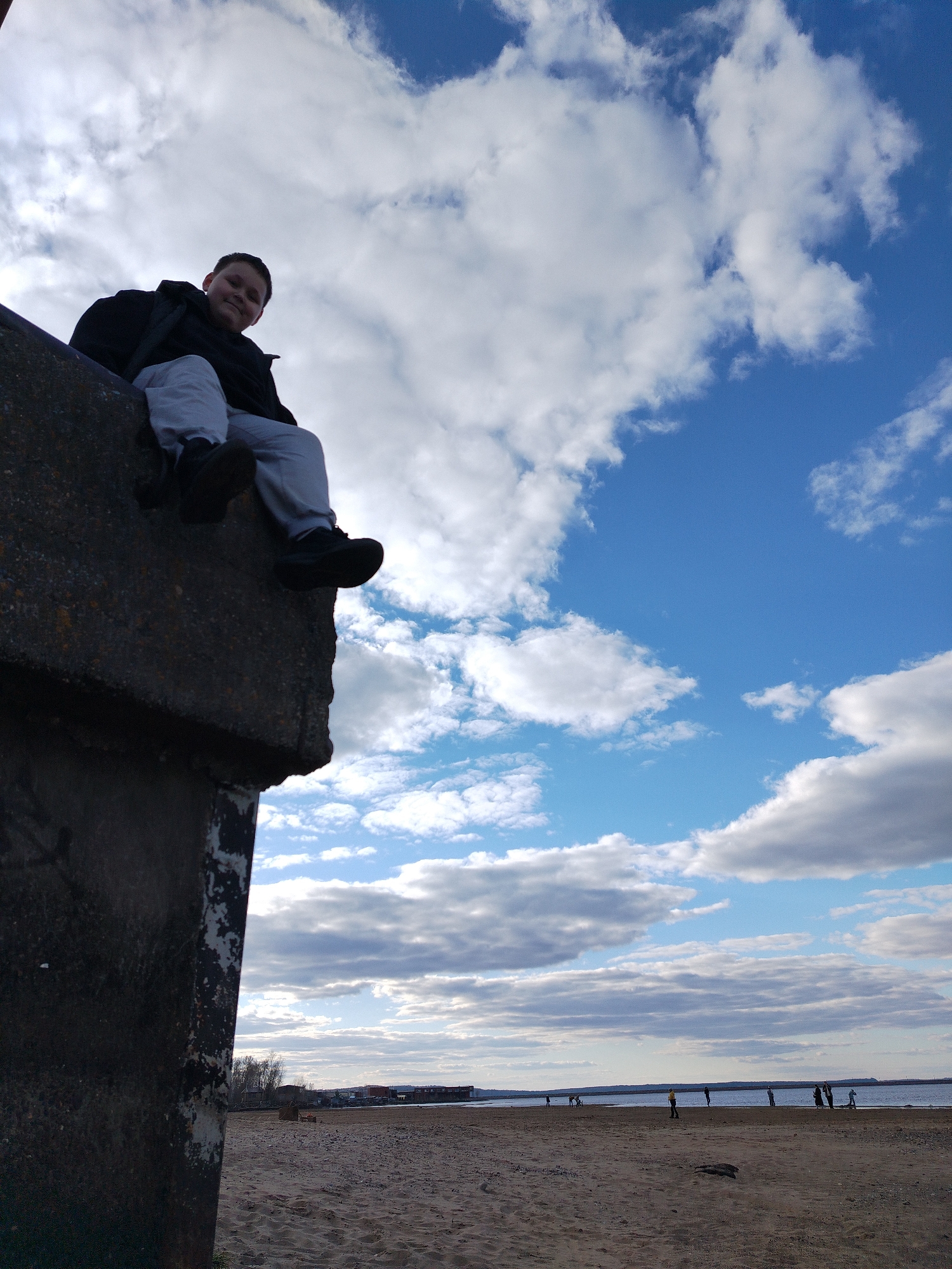 A child sits on the edge of a concrete structure overlooking a sandy beach in Unknown, Unknown. The child is wearing a dark jacket, light-colored pants, and dark shoes. The sky is blue with white clouds. In the distance, there is water, and several people are standing on the beach.