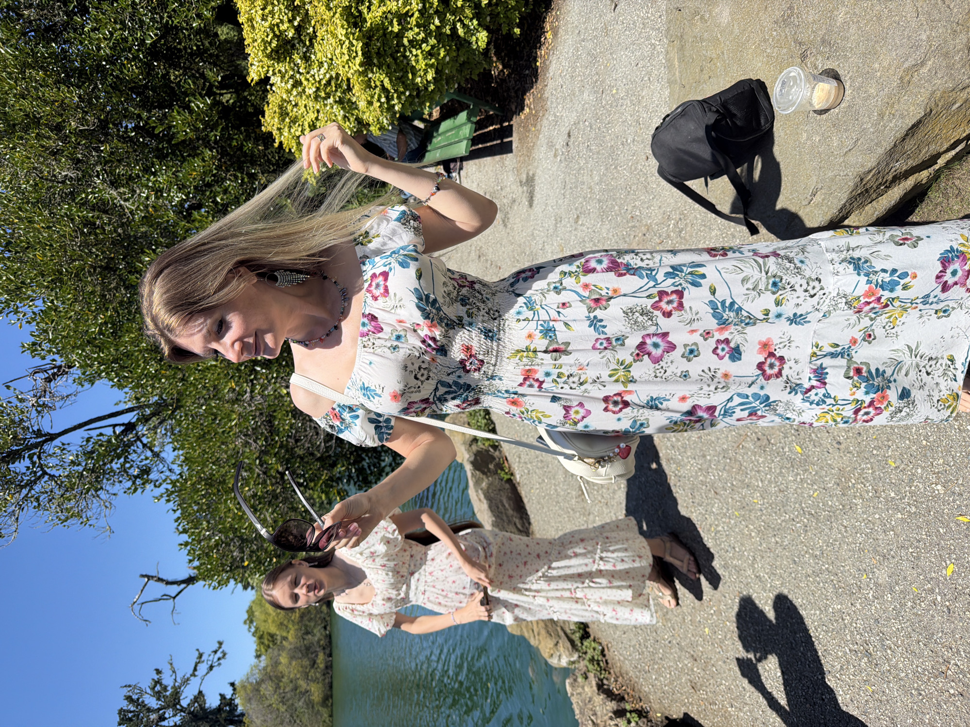 Two women are standing near a body of water on a sunny day. The woman in the foreground, wearing a floral print dress, is holding sunglasses and looking to the left. She has blonde hair and is wearing a necklace and earrings. A white bag is slung over her shoulder. To her right, a black backpack and a disposable cup with a drink are placed on a rock.
Behind her and to the left, another woman, wearing a similar floral print dress, stands closer to the water's edge. She has long brown hair and is looking towards the water. Her hands are resting on her dress.
The environment appears to be a park or garden, with lush green trees and bushes in the background. The body of water is calm and reflects the blue sky. The ground is paved with asphalt. The presence of a green park bench in the background suggests a public outdoor space.
Given the lush greenery, the presence of a large body of water, and the sunny weather, Golden Gate Park in San Francisco, United States of America, is a likely location. The specific area might be near Stow Lake or a similar water feature within the park.