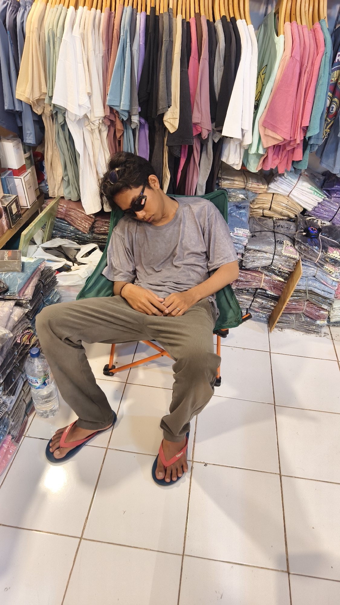 A medium, full shot depicts a man asleep in a chair inside a retail clothing store in Pecatu, Indonesia. The man is wearing sunglasses, a gray t-shirt, and olive-colored pants. He is seated in a green folding chair with an orange frame. His feet are clad in flip-flops, the left one being blue with a pink strap and the right one red with a blue strap.
Behind him, numerous shirts on hangers form a wall of varied pastel colors. Stacks of folded clothing are visible to his right and left. A water bottle and some boxes of goods rest near him on the left. The floor is tiled in light, neutral squares. The general environment suggests a small, locally-owned business.