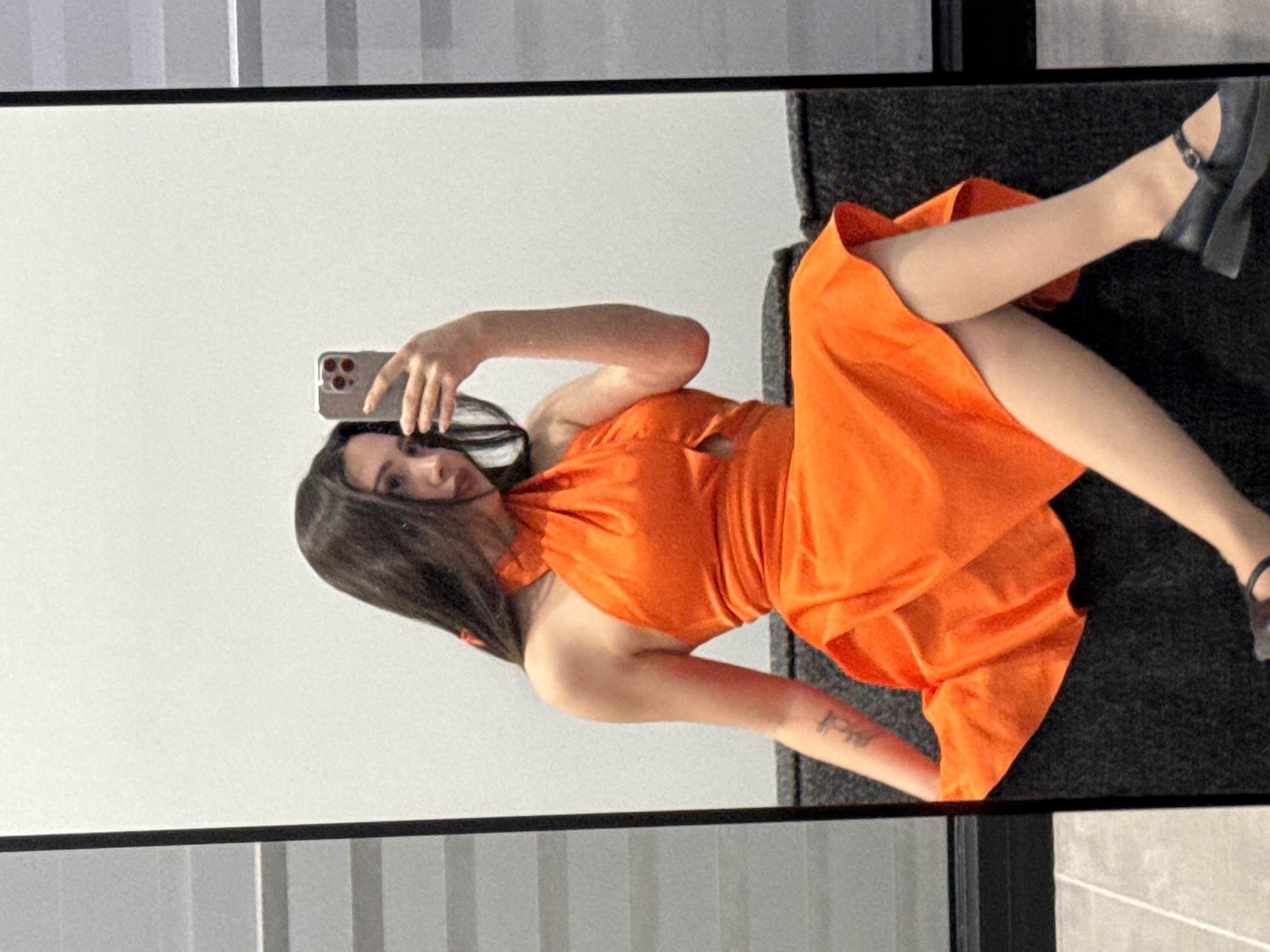 This image, taken in Yerevan, Armenia, captures a young woman taking a mirror selfie indoors. The woman, with long dark hair, is reflected in a large, dark-framed rectangular mirror. She is wearing a vibrant, bright orange, sleeveless or halter-neck dress with a flowing skirt. Her legs are crossed, and she has on black chunky high heels with an ankle strap.
She holds a smartphone with multiple camera lenses (resembling an iPhone) in her right hand, aiming it towards her reflection. Her left arm is bent, resting near her side. A tattoo is visible on her right forearm, though the text is illegible. Her expression is neutral as she looks towards the phone.
The background visible in the mirror is a light-colored wall with subtle horizontal gray lines in the upper section, possibly a window or architectural detail. The foreground area, where the woman is presumably positioned, appears to be a dark, textured surface, possibly carpet or upholstery. The setting suggests a modern indoor environment. The image is oriented with a 90-degree counter-clockwise rotation from its original capture, making the subject appear horizontal within the frame.
