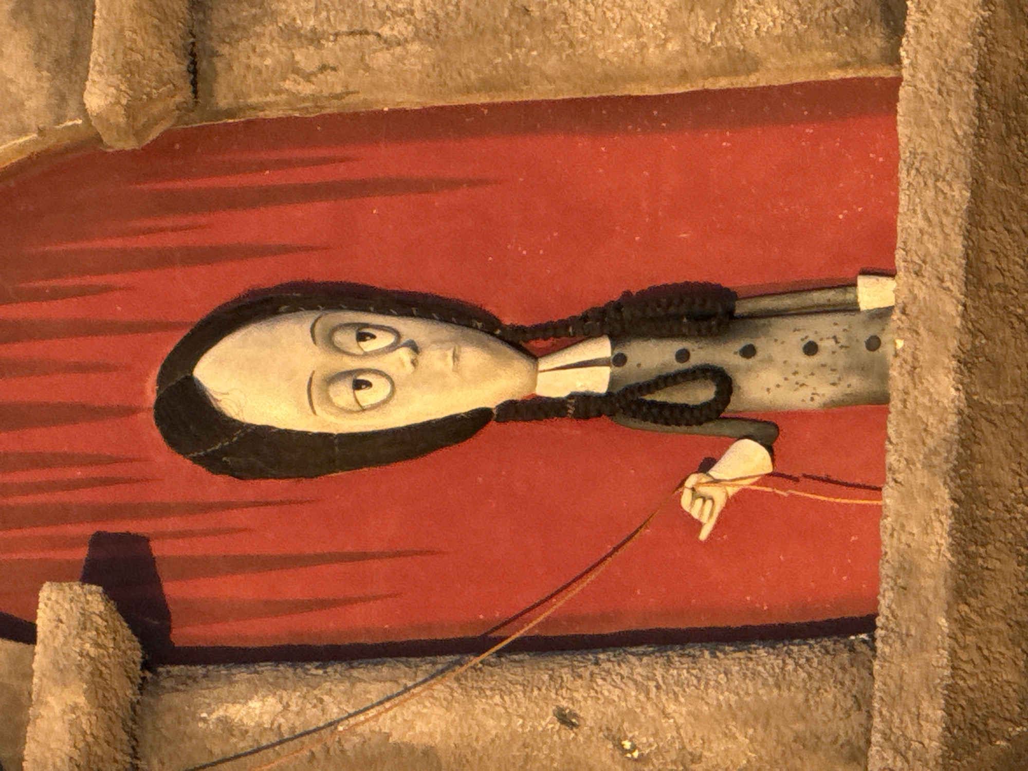 The image shows a stop-motion animation puppet resembling Wednesday Addams. She has a pale, elongated face with large, round eyes and a small mouth. Her black hair is pulled back into two braids. She is wearing a dark dress with a white collar and several black buttons. Her right hand is extended, holding a thin string. The puppet is positioned in front of a red backdrop with vertical lines, suggesting a theatrical setting. The scene is framed by textured, stone-like architectural elements. The image is a close-up and does not provide any geographical context.