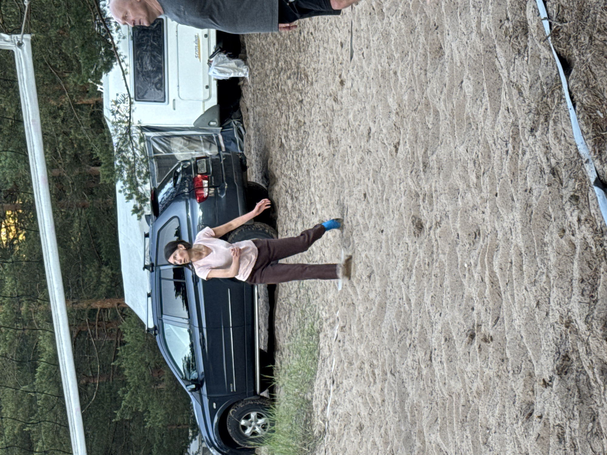 A woman in a light pink t-shirt and brown pants is captured mid-action in a sandy area, possibly playing volleyball, with a dark SUV and a white camper trailer visible in the background, amidst a backdrop of pine trees. Another man, wearing a grey t-shirt and black shorts, stands to the right, looking away from the camera. The ground is sandy and uneven, with a volleyball net strung across the top of the frame. The scene appears to be a campsite, potentially located in or near Ozerki (Krasnodolinskaya), Russia, given the presence of the camper and the natural environment.