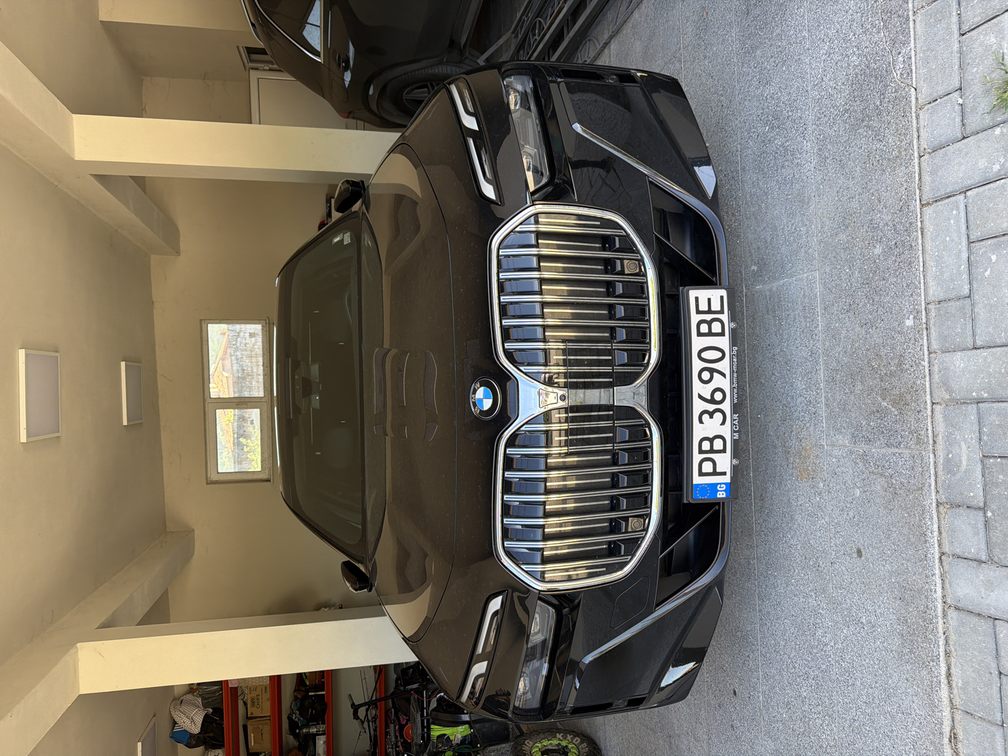 A black BMW with Bulgarian license plate PB 3690 BE is parked nose-in in a garage. The garage is attached to a building with tan walls and a small window. Various items are stored in the garage, including a green four-wheeled vehicle and shelving units filled with boxes. The car is partially parked on a paved driveway. The location is Asenovgrad, Bulgaria (as indicated by the license plate).