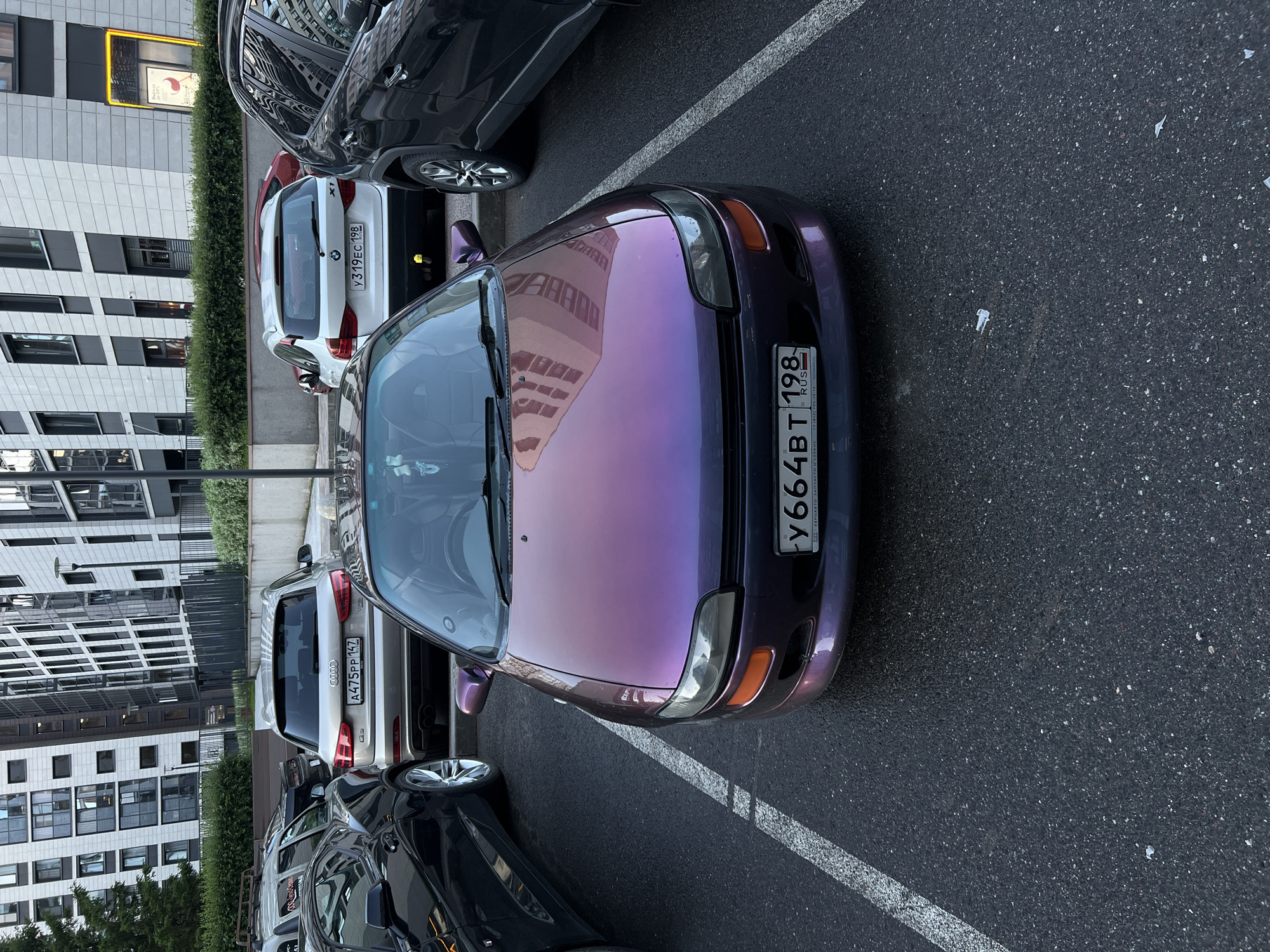 In an outdoor parking lot with asphalt pavement, multiple cars are parked. In the foreground, a purple iridescent hatchback is parked, facing the camera. It has a license plate that reads "y664BT 198". To the left of the purple car is a dark-colored coupe. To the right of the purple car is a black SUV. Behind the purple car, a silver Audi SUV is parked. Further back and to the right of the Audi, a white BMW SUV is parked. The background features modern multi-story buildings with glass facades. The sky is overcast. The scene appears to be in an urban residential area in Russia.