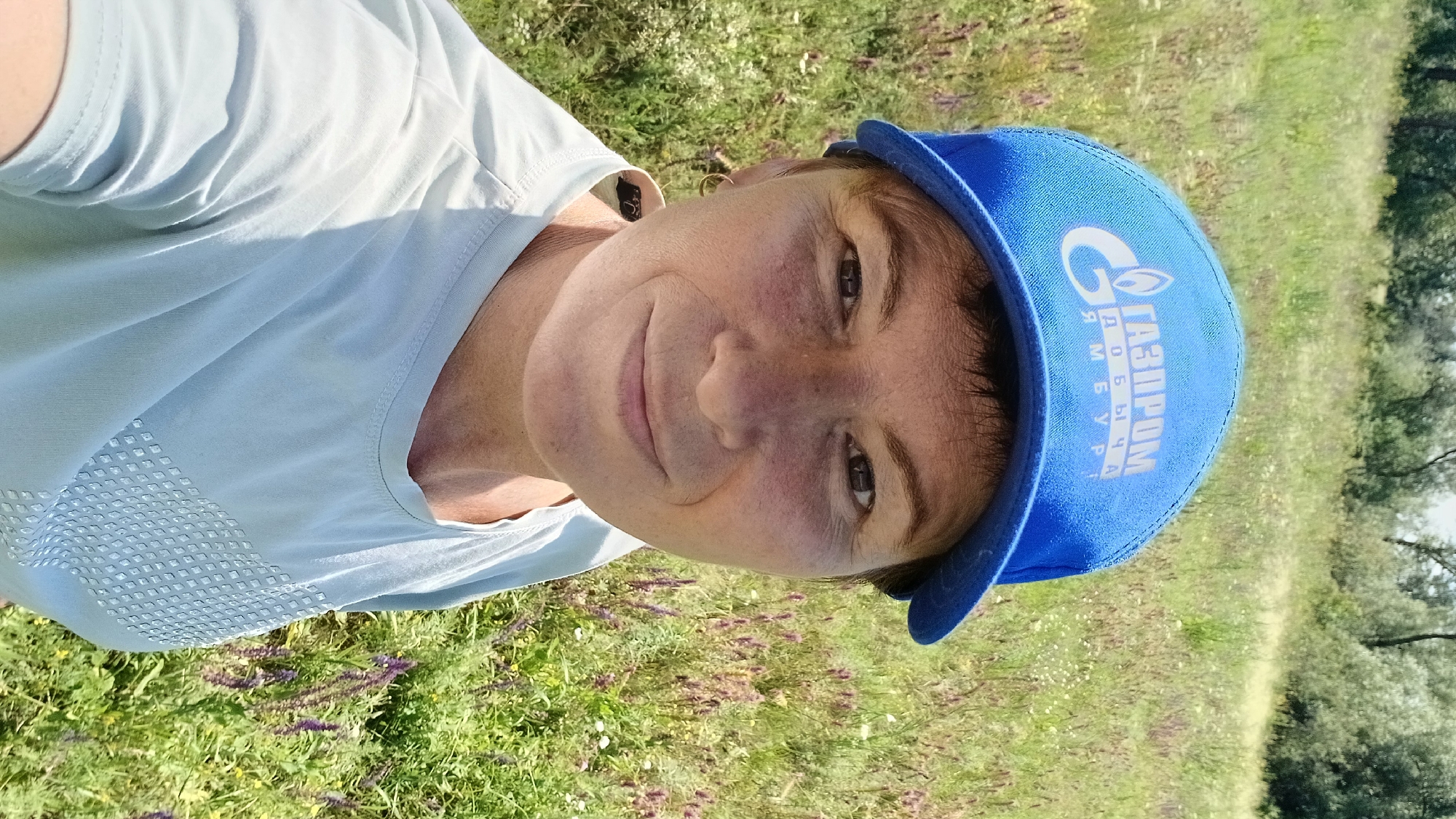 A woman wearing a blue baseball cap with the "Gazprom" logo and a light blue t-shirt takes a selfie outdoors in a grassy field with trees in the background. The woman has short brown hair and is smiling. The image appears to have been taken in Nadezhdenka, Russia.