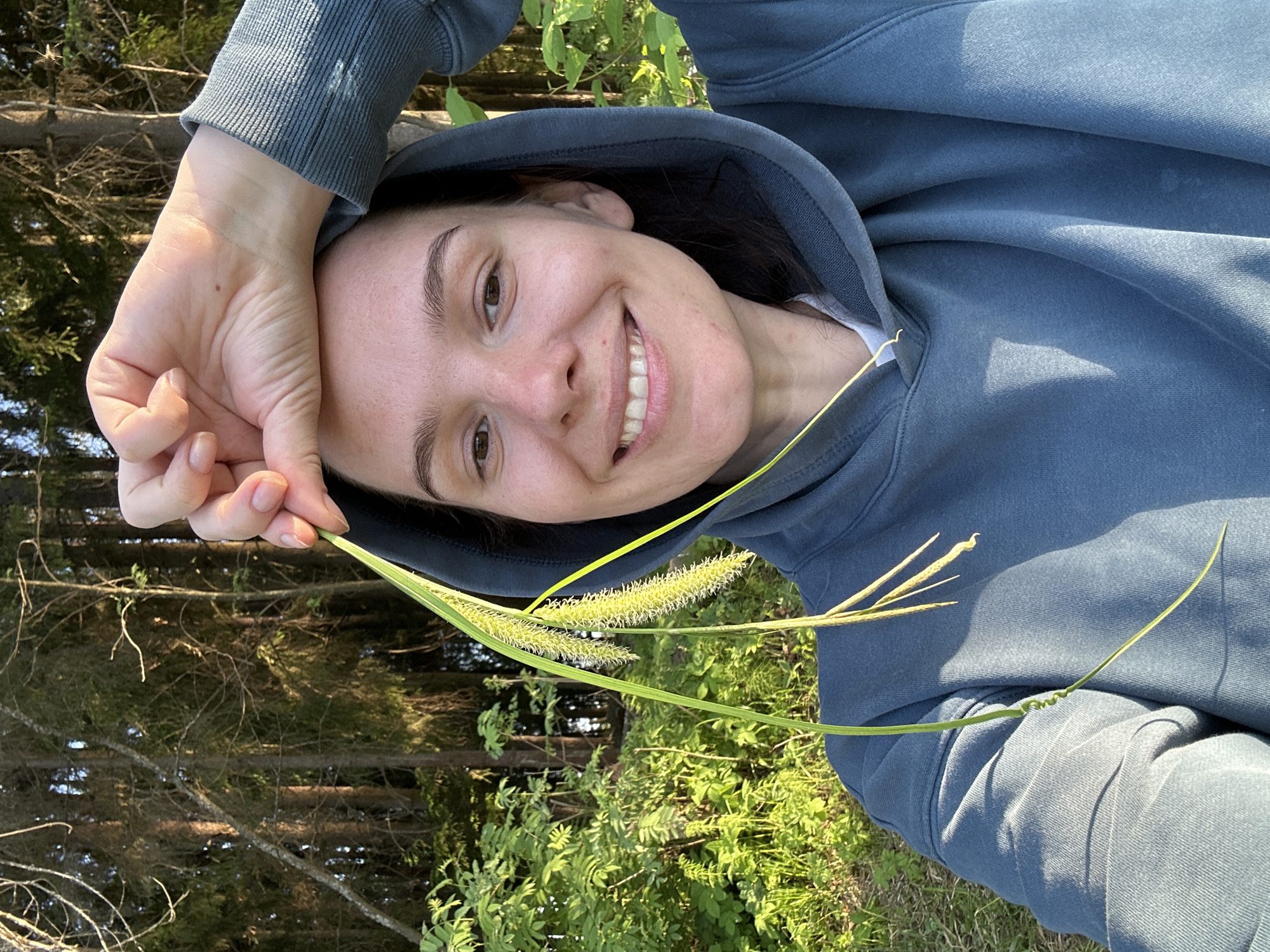 A young woman is photographed outdoors in Bormist, Russia. She is holding a piece of grass with seed heads close to the camera, smiling. She is wearing a blue hooded sweatshirt. The background shows a blurred view of trees and green foliage. The scene is lit with natural daylight.