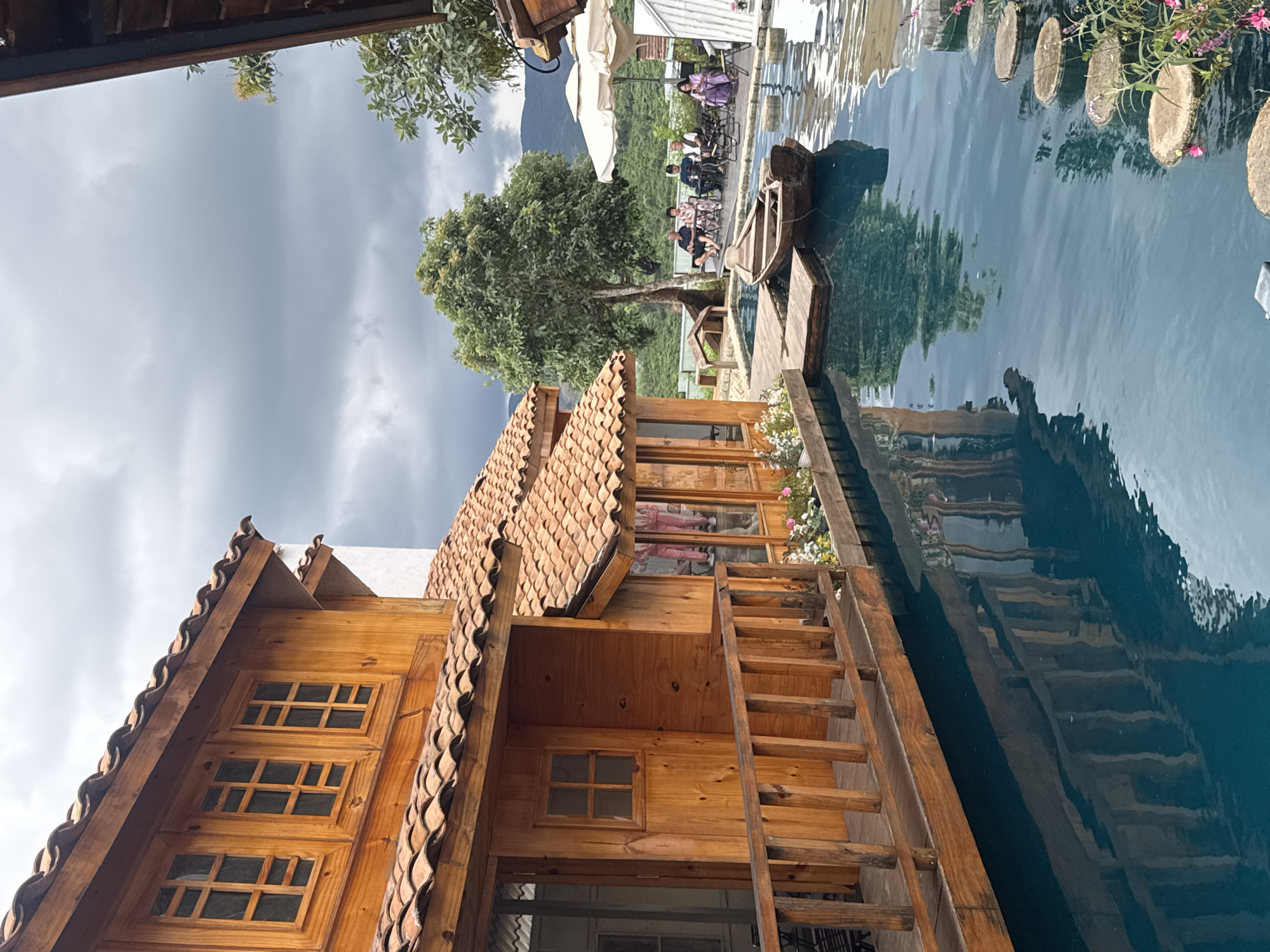 This is an exterior, eye-level, medium shot of a waterfront property in Thôn 3, Vietnam. The image features a two-story wooden building with a terracotta tile roof, possibly a guesthouse or restaurant, situated on the edge of a calm, blue body of water. A wooden balcony with a railing extends from the building over the water. In the foreground, a wooden rowboat is moored to a small wooden dock. A pathway of stepping stones leads across the water towards the right side of the image, where several people are seated at tables under umbrellas, enjoying the view. Lush green trees and rolling hills form the background. The sky is overcast with dark clouds. The overall scene suggests a serene, relaxing atmosphere.