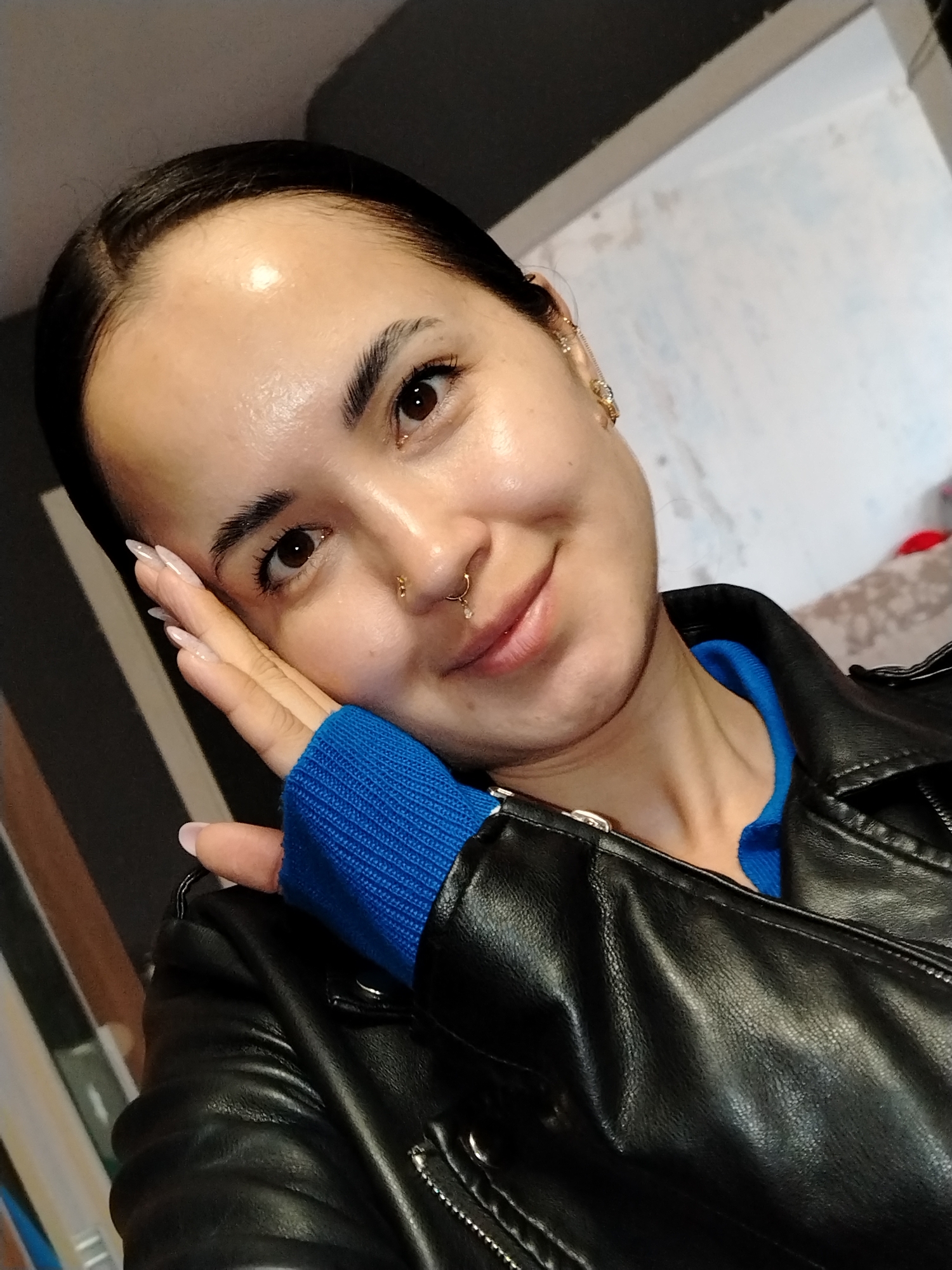A person, likely female, is centered in a selfie-style shot. She is wearing a black leather jacket over a blue garment. Her face is tilted slightly to the right, with her left hand near her face. She has dark hair pulled back and various piercings in her ear and nose. The background consists of a gray wall, a white wall, and a glimpse of some furniture. The location in the city of Noviy, Russia is unknown.