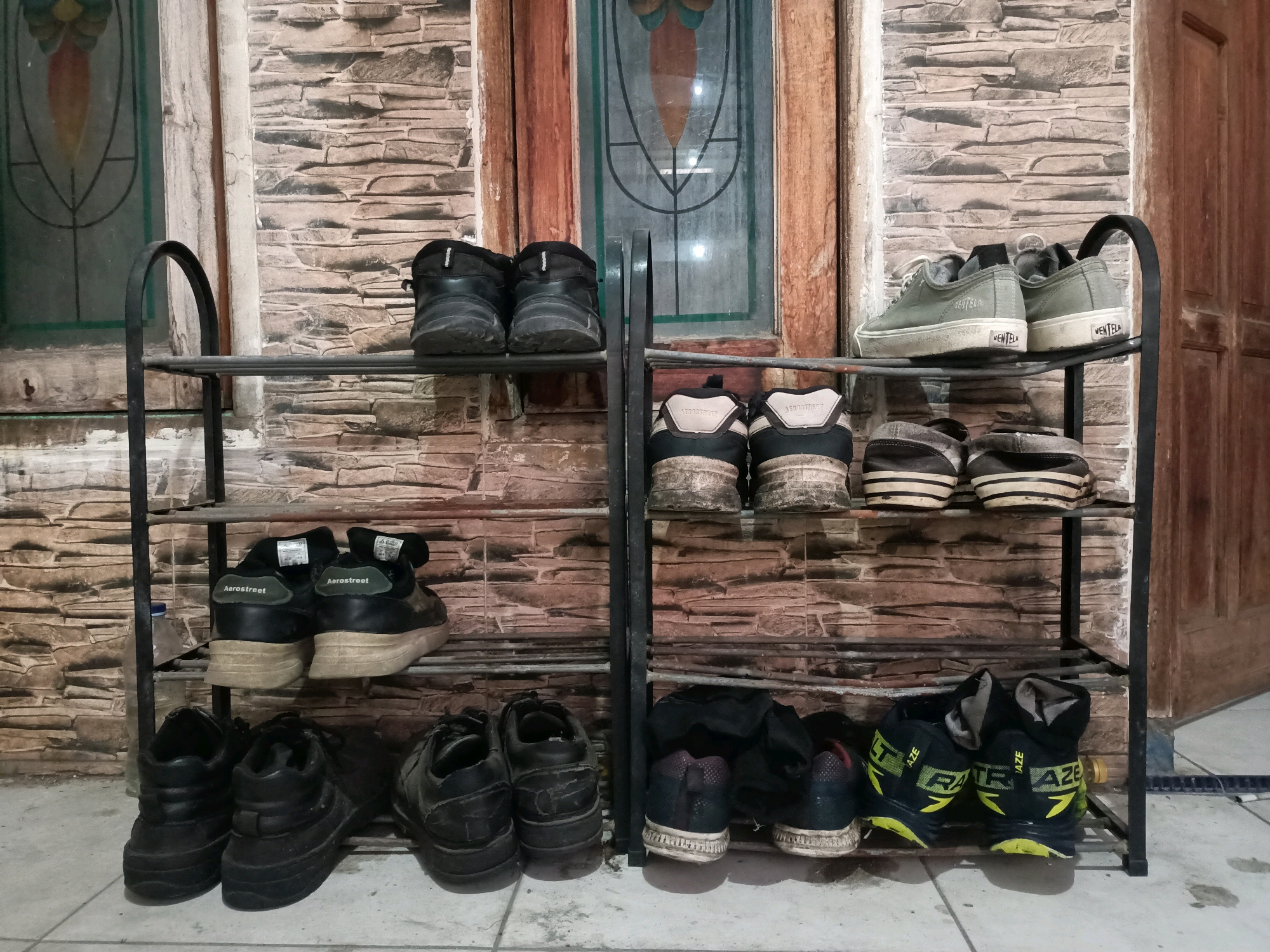 In Ngargosari, Indonesia, a two-tiered metal shoe rack stands against a textured stone wall, positioned in front of a wooden door with stained glass detailing. The rack holds several pairs of footwear. On the top shelf of the left side, two black boots are visible. Below them, on the second shelf, two black sneakers with white accents are arranged. The bottom shelf on the left features four black shoes, including two pairs of low-top sneakers and two pairs of chunky-soled boots.
The right side of the shoe rack has a matching two-tiered structure. The top shelf displays two pairs of low-top canvas sneakers, one pair in a light grey and another in a darker grey. The second shelf from the top holds two pairs of dark-colored sneakers with striped soles. The bottom shelf on the right is occupied by two pairs of black athletic shoes with yellow accents and visible branding. The overall scene depicts an organized storage of shoes in an outdoor or semi-outdoor setting.