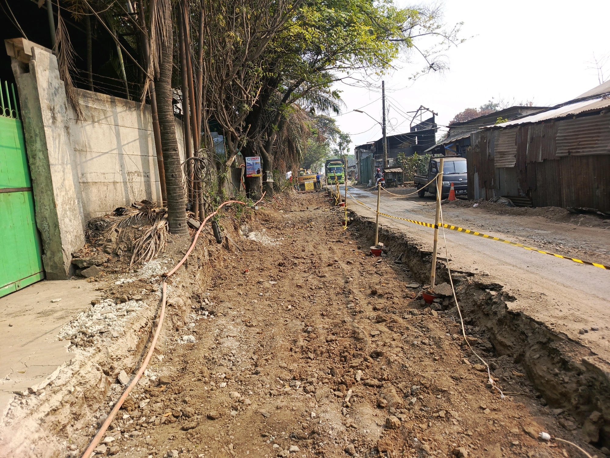 This image depicts a street under construction in Tangerang, Indonesia. The scene is dominated by a deep excavation along the side of the road, filled with loose dirt and debris. A pink hose snakes through the trench. The road surface itself is unpaved and appears to be composed of dirt and gravel. Yellow and black caution tape is strung along wooden poles, marking off the construction area.
In the background, construction equipment, including a yellow Komatsu machine, is visible. Several vehicles are present, including a green truck, a black pickup truck, and a motorcycle. Structures line the right side of the road, appearing to be makeshift or dilapidated buildings made of corrugated metal. Vegetation, including trees with abundant foliage and palm trees, frames the left side of the scene, with utility poles and wires running overhead. A concrete wall and a green gate are also visible on the far left. The sky is bright and overcast.