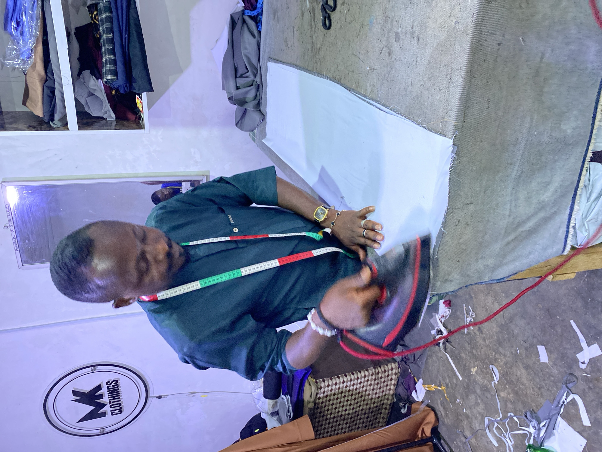 In a brightly lit indoor workshop in Jos, Nigeria, a man is actively engaged in the process of ironing fabric. The man, likely a tailor, is positioned in the foreground, wearing a dark green or blue shirt with a measuring tape draped around his neck. On his left wrist, he wears a gold watch and a black and white bracelet. His right hand holds a dark iron with a red cord, pressing down on a large piece of light blue fabric laid out on a textured, grey work surface, which appears to be a cutting or ironing table.
In the background and to the left, various garments and fabrics are visible, some hanging on racks, others folded. A bright light source is reflected in a mirror on the left wall, above which some partial text, possibly "Bouliiz x St...", can be seen. Below the mirror, a round, illuminated sign prominently displays the text "MK CLOTHINGS". The floor, made of concrete or a similar material, is littered with fabric scraps, indicating an active workspace. The man appears focused on his task, contributing to the professional and industrious atmosphere of the tailoring shop.
