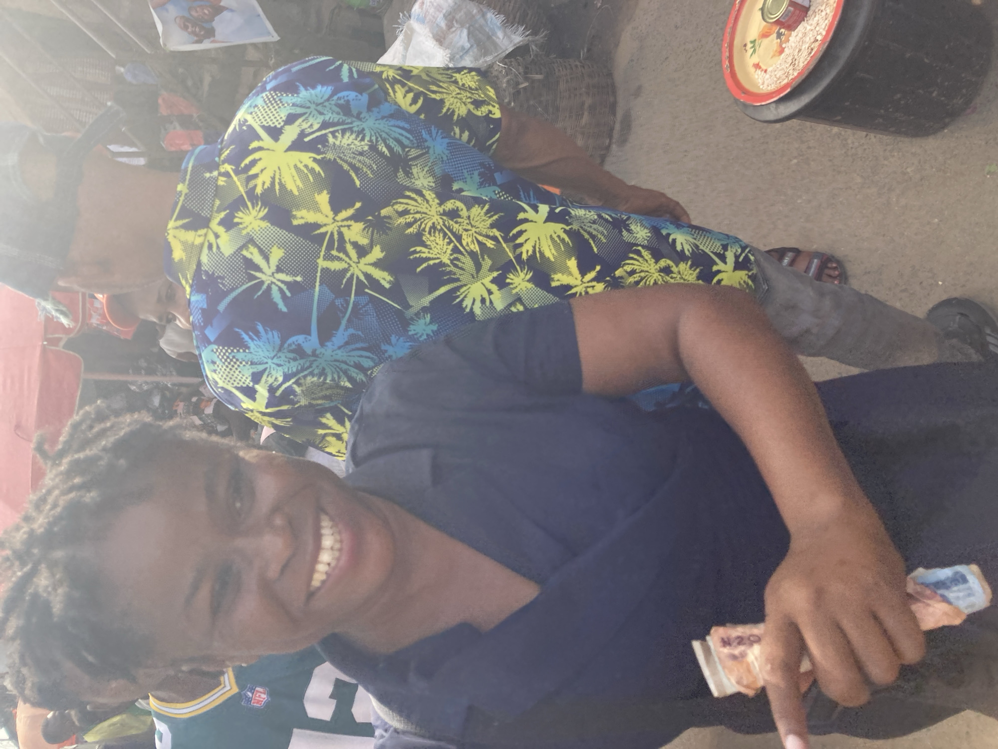 The image shows two individuals in what appears to be an outdoor market in Ura Nta Umuarandu, Nigeria. The individual in the foreground is smiling and holding what looks like Nigerian currency. This person is wearing a dark blue shirt over a Green Bay Packers jersey. The second person, behind, is wearing a short-sleeved shirt with a palm tree pattern. A large black container is visible, likely holding goods for sale. The environment seems busy, with other people and items partially visible in the background.