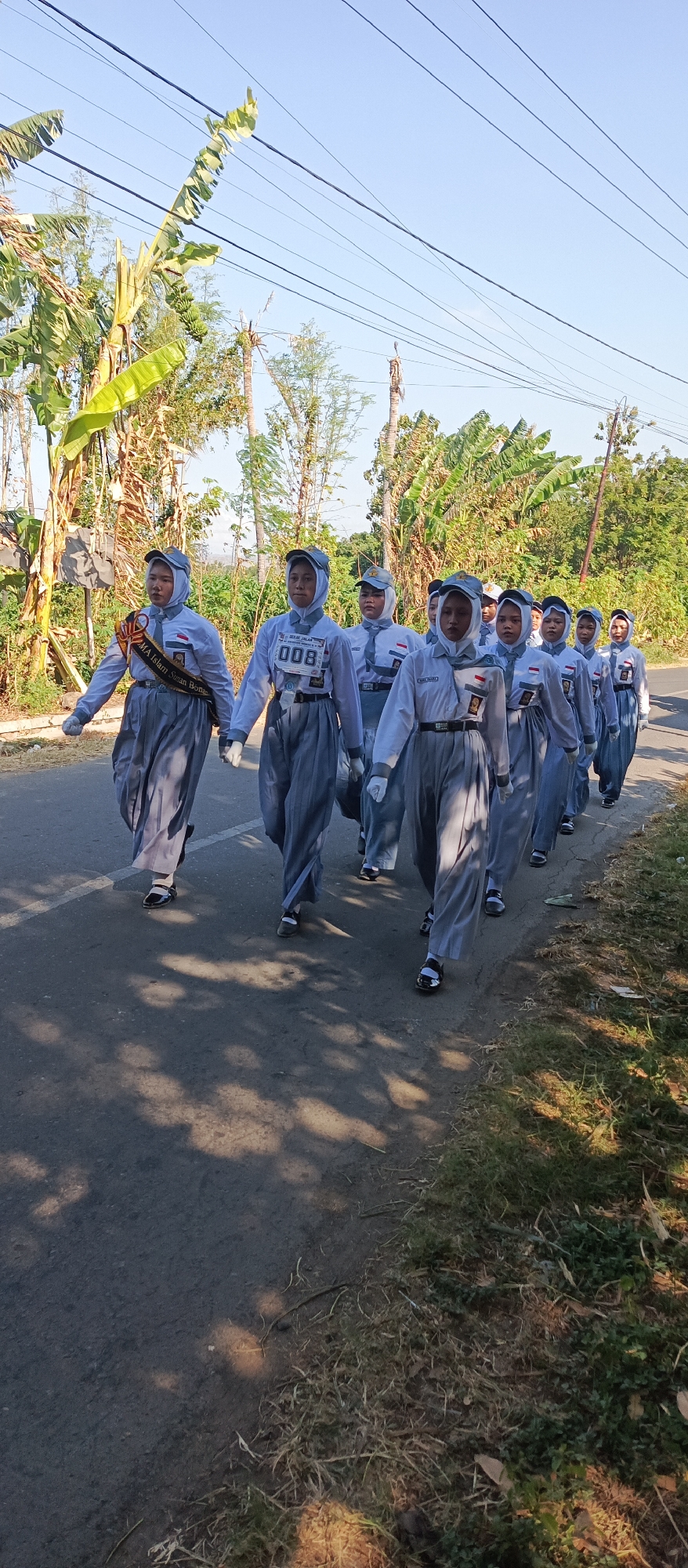 In Ketowan, Indonesia, a group of young women wearing matching grey uniforms and white hijabs are marching in single file on an asphalt road. They are participating in a parade, indicated by a sash worn by the lead participant that reads "MA Islam Sumar Bon...". The uniform consists of a long-sleeved white top with grey accents and a grey skirt or trousers. They are also wearing grey caps and white gloves. Some participants have badges or numbers displayed on their uniforms. The environment is outdoors, with lush green vegetation and trees lining the side of the road. Banana trees are prominent in the background, and a power line runs across the top of the image. The sky is clear and blue, suggesting it is a sunny day. The overall scene depicts a formal procession in a rural or semi-rural Indonesian setting.