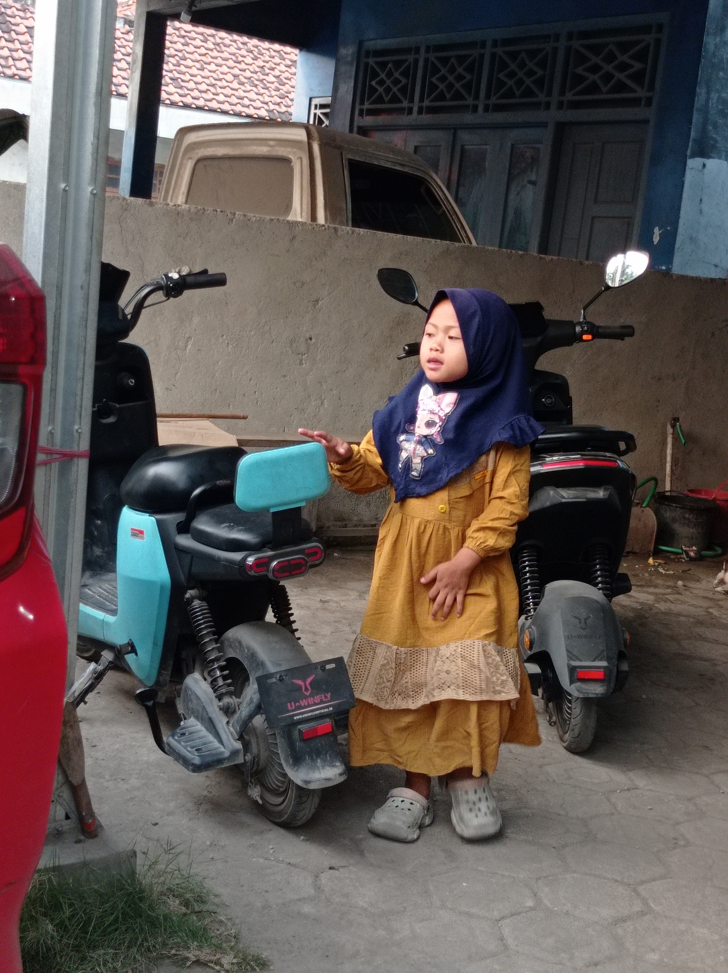 A young girl wearing a dark blue hijab and a yellow dress stands next to a light blue electric scooter. The girl is looking to her left, with her right hand outstretched towards the scooter. Her left hand rests on the seat of the scooter. The scooter has a black seat and a light blue body. Behind the scooter, a dark gray vehicle, possibly a truck, is partially visible. In the background, a building with a tiled roof and blue walls is seen. The scene appears to be an outdoor area, possibly a driveway or yard, with a paved ground surface. There is some grass to the left of the scooter. The overall lighting suggests it is daytime. The context of Maribaya, Indonesia is not directly observable from the visual elements of the image.
