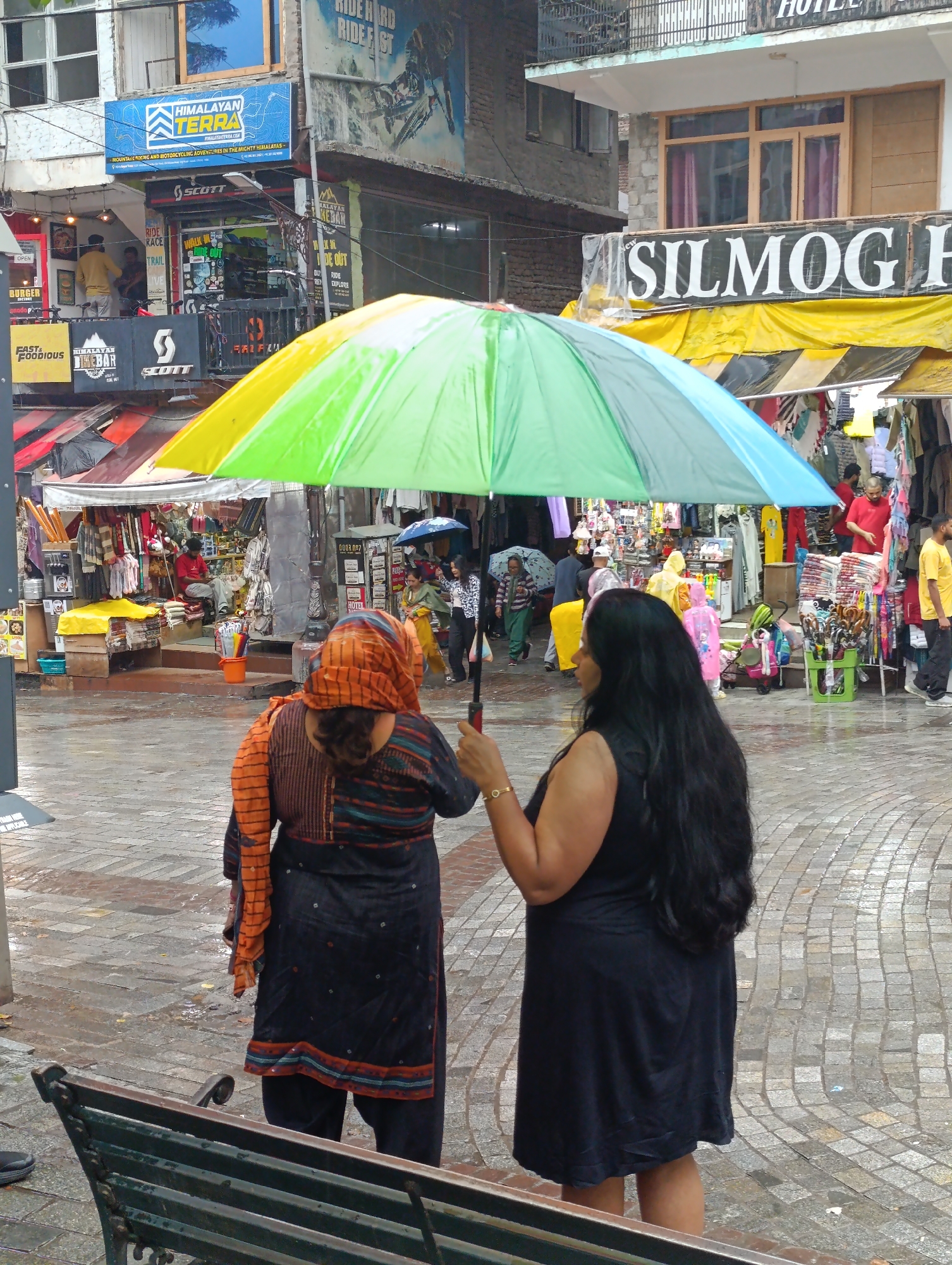 In Manali, India, two women stand under a large, multi-colored umbrella on a wet cobblestone street. The woman on the left, seen from behind, wears a dark outfit with an orange and striped headscarf, and an orange shawl draped over her shoulders. The woman on the right, also seen from behind, has long black hair and wears a black sleeveless dress. In the background, a marketplace bustles with activity, with stalls selling clothing and other goods. A neon sign for "HIMALAYAN TERRA" is visible on the left side of the image, indicating the location. The overall scene depicts a typical day in a busy Indian street market.