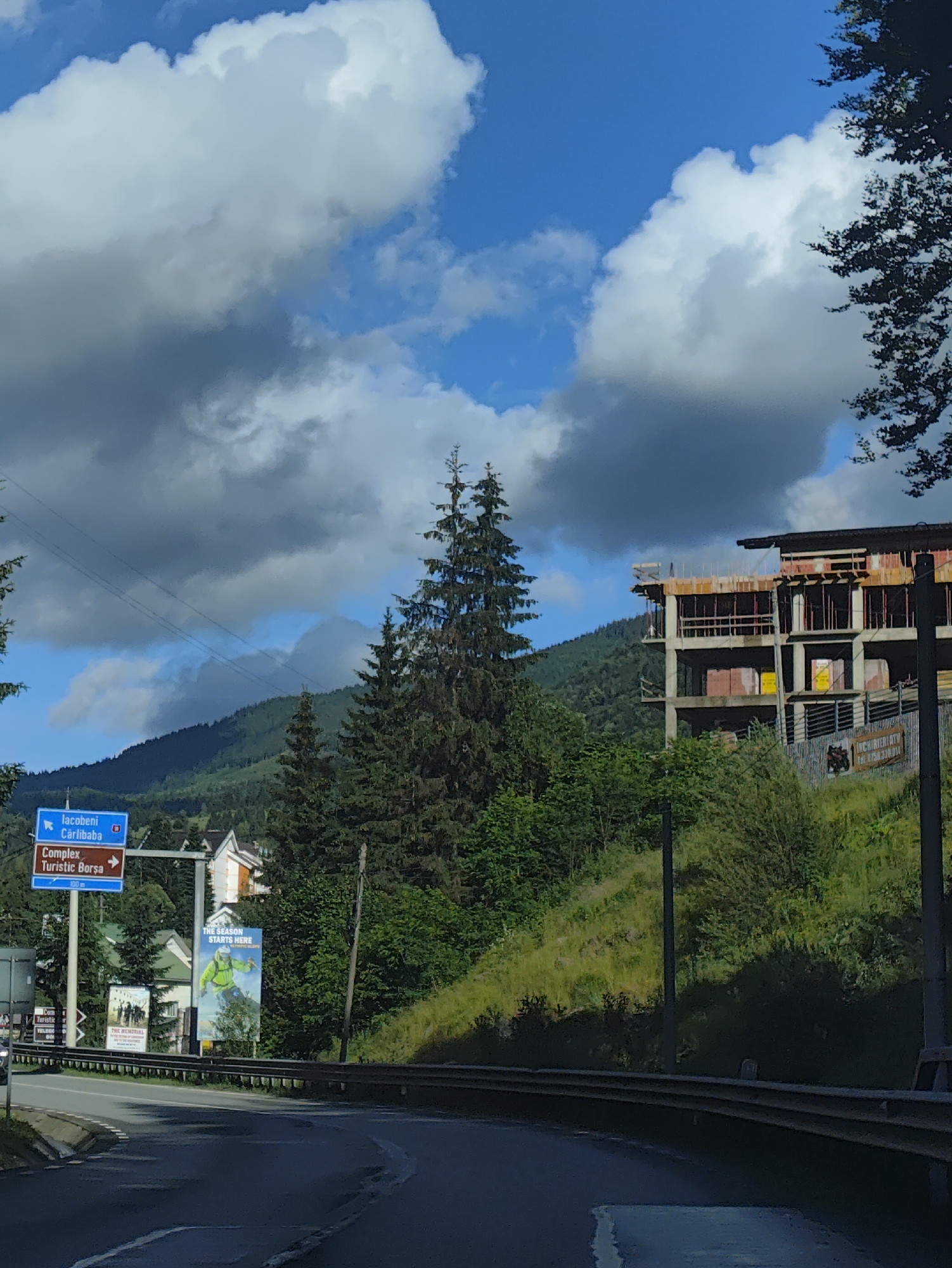 This media file depicts a mountainous landscape in Borșa, Romania, on a partly cloudy day. In the foreground, a wet asphalt road curves to the right, bordered by a metal guardrail. To the left of the road, a blue directional sign points left towards "Iacobeni" and "Cărlibaba" and then right towards "Complex Turistic Borșa." Next to this sign, a large billboard displays a person in a bright green ski suit, with the text "THE SEASON STARTS HERE" at the top.
Further to the left, several houses with pitched roofs are visible nestled amongst trees. A second, smaller billboard with black and white imagery is also present. The middle ground is dominated by a steep, grassy hill dotted with trees. Atop this hill, a multi-story building is under construction, with visible concrete floors, scaffolding, and some yellow signage. The structure appears to be a large hotel or resort.
In the background, lush green mountains rise, partially obscured by dramatic, dark grey and white clouds against a bright blue sky. Tall evergreen trees are scattered throughout the scene, particularly on the left and right sides. The overall impression is one of a developing tourist area in a natural, scenic setting. The construction of the building suggests ongoing development for tourism, likely related to the nearby ski resort, as indicated by the billboard. The weather appears to be dynamic, with both sunshine and heavy clouds present.