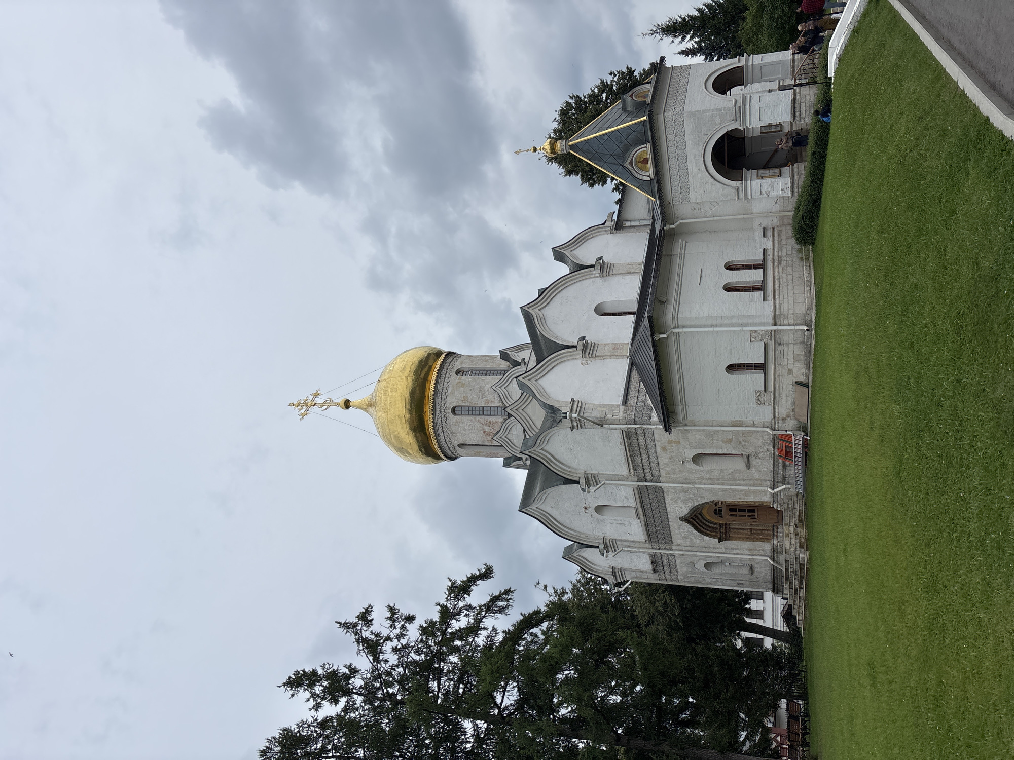 This is a frontal, eye-level, full shot of the Dormition Cathedral in Zvenigorod, Russia, under an overcast sky. The cathedral is whitewashed with a large, golden dome topped with a cross. Adjacent to the main dome is a smaller, dark-tiled roof with a golden dome and cross. The cathedral has arched windows and decorative friezes. To the left of the cathedral are tall evergreen trees. In front of the cathedral is a well-maintained lawn and a paved walkway. Several people are visible near the entrance of the cathedral, some standing and others walking. A ladder and some orange plastic barriers are placed near the entrance on the right side.