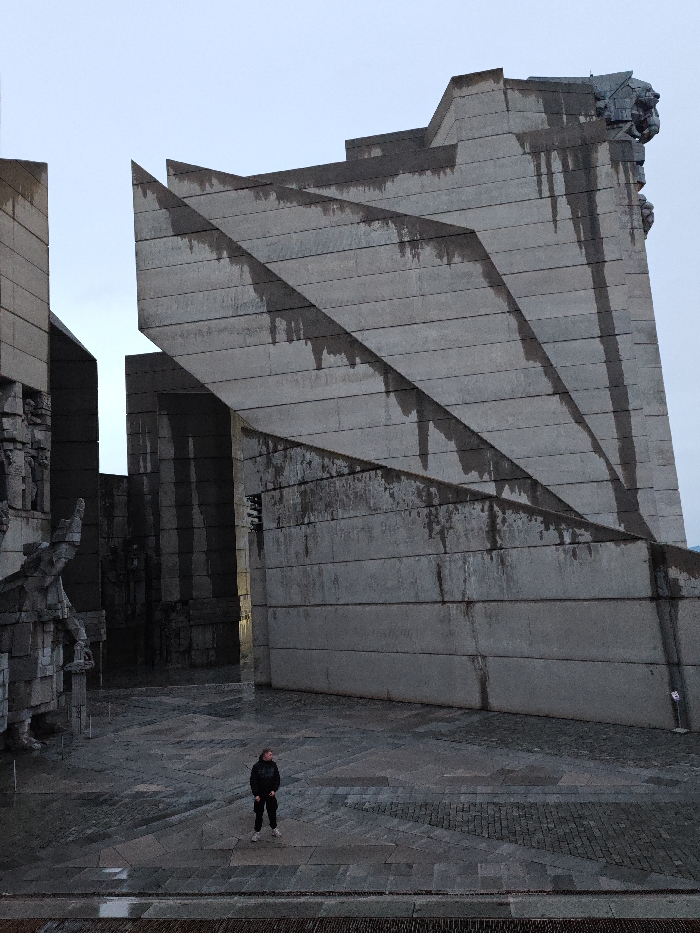 The image displays the Monument to 1300 Years of Bulgaria in Shumen, Bulgaria, under an overcast sky. The foreground consists of a dark grey, wet paved surface with geometric patterns. A single individual stands centrally, facing slightly left. This person wears a dark hooded jacket, dark trousers, and light-colored athletic shoes, standing still and observing the monumental structures.
The monument itself features large, angular concrete blocks in a brutalist architectural style. A prominent stepped concrete wall on the right exhibits dark vertical streaks from water runoff and weathering. Its top-right edge incorporates a large, stylized sculptural relief, depicting a fierce head. To the left, interconnected concrete structures present multiple integrated, dark stone-carved figures, partially visible. The scene is illuminated by diffuse light.