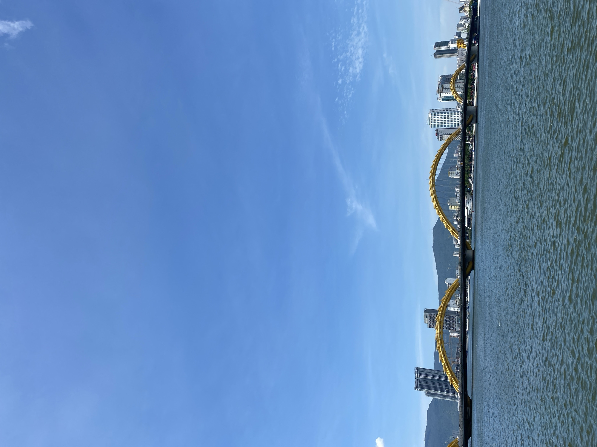 This image captures the Dragon Bridge, a distinctive landmark in Đà Nẵng, Vietnam, spanning the Han River. The bridge, painted in a vibrant yellow, features a dragon-like structure arching over the roadway. In the background, a modern cityscape rises with several tall buildings, including skyscrapers, and a range of hills or mountains. The Han River flows in the foreground, appearing calm with ripples on its surface. The sky is a clear blue, with a few wispy white clouds scattered across it, indicating a bright, sunny day. The scene is devoid of people or any specific actions, presenting a static, architectural and environmental view of the city.