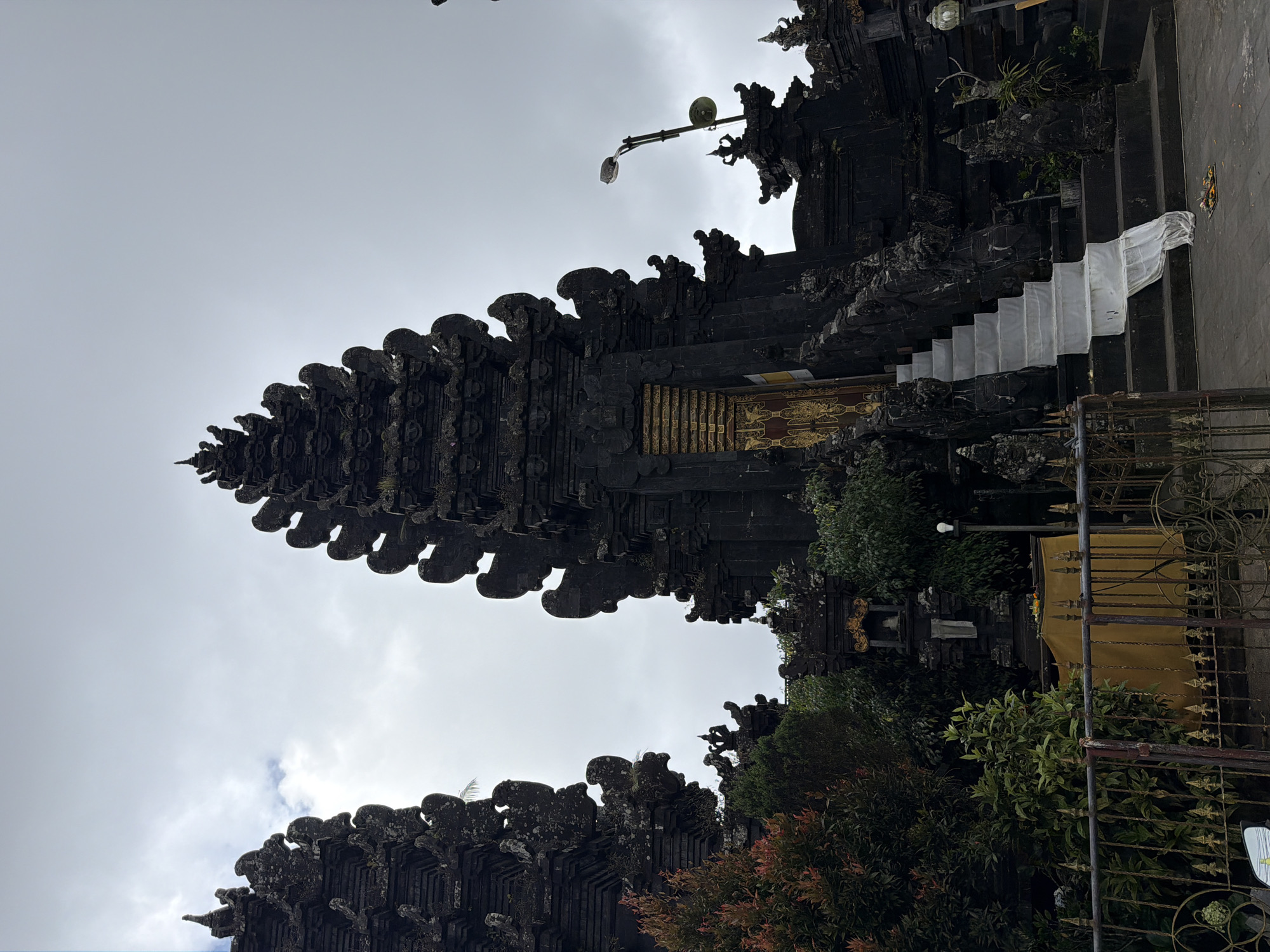 The image captures a view of a traditional Balinese temple entrance, oriented vertically. The temple is constructed from dark stone, intricately carved with decorative elements characteristic of Balinese architecture, such as tiered roofs and ornate sculptures. A prominent feature is the towering gateway, which resembles a Candi Bentar or a split gate, a common sight in Bali.
In the foreground and to the right, there is lush greenery, including trees and shrubs, which partially obscure the view of the temple. A yellow tarp is visible behind a metal fence, suggesting some sort of temporary covering or decoration. To the left of the temple gateway, a set of white-covered stairs leads upwards, suggesting an elevated pathway or an entrance to a higher level of the temple complex.
The sky above is overcast with clouds, indicating a possibly cloudy or impending rainy weather. A street lamp with a light bulb is mounted on a pole extending from the temple structure, suggesting it is a functional part of the temple grounds, possibly for evening illumination. There is no visible text in the image. The overall mood is somber due to the overcast sky and dark architecture, yet it also conveys a sense of historical and cultural significance associated with the temple. The scene appears to be set in a location like Kintamani, Indonesia, known for its temples and natural beauty. No people are visible in the frame.