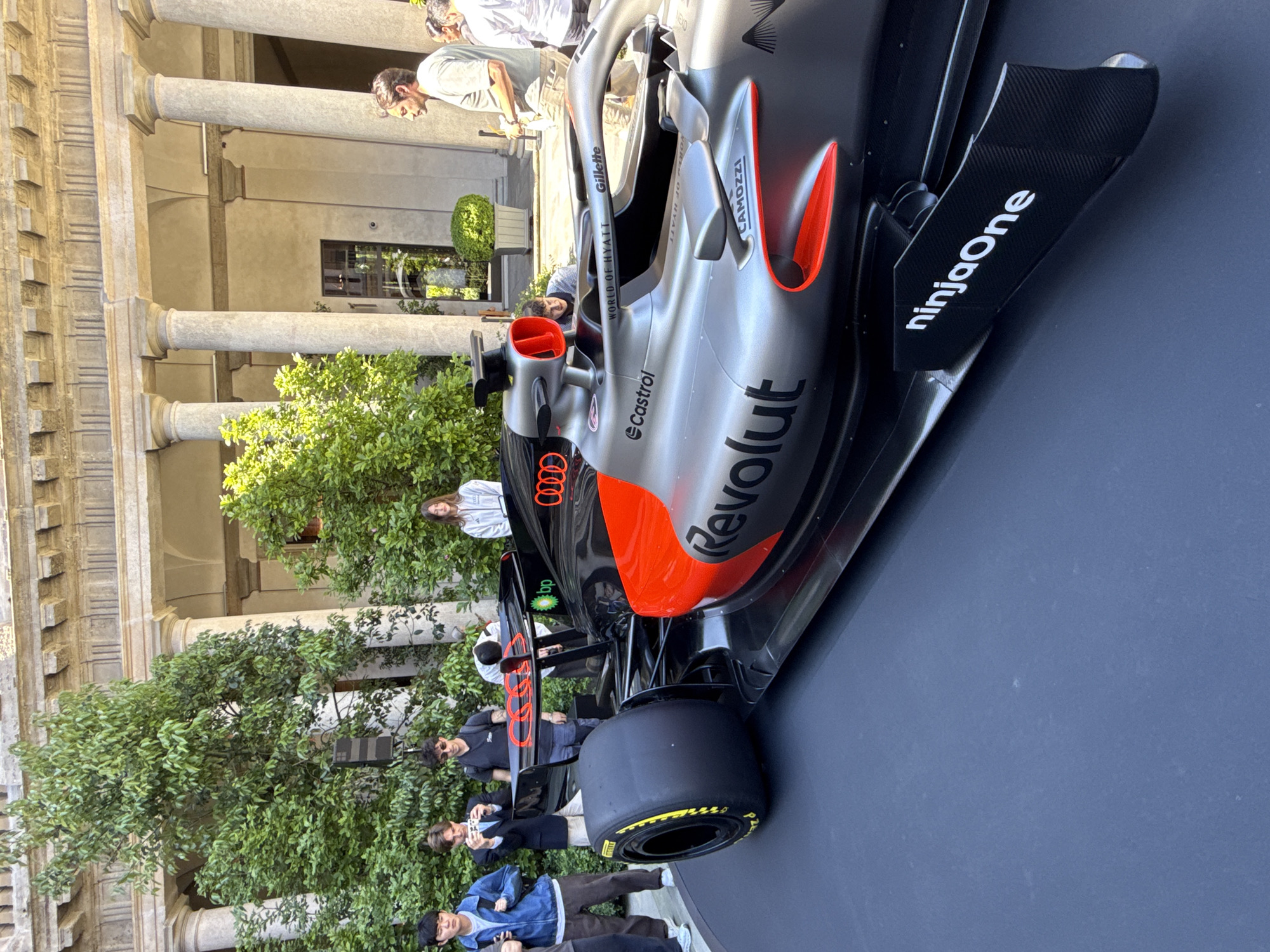 The image shows an Audi Formula One car on display. The car is predominantly silver and black with orange accents and sponsor logos including Revolut, Castrol, and NinjaOne. Several individuals are present, including people inspecting the car and taking photos. The car is located in a courtyard with classical columns, suggesting an architectural or historical setting. Given the prompt's instruction, it is in Milan, Italy.