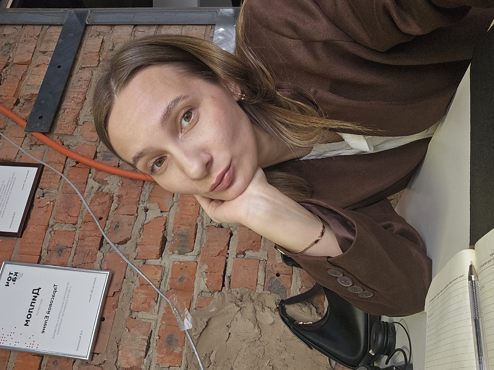 The image depicts a woman resting her head on her hand while posing against a brick wall. She wears a brown blazer over a white top and has a gold bracelet and earrings. In the background, there are framed certificates in Russian. The lower certificate features a logo of the letters "Kg" over "MOT". Next to her is an open notebook and a pen. A black bag and headphones are placed beside the notebook. An orange cable runs along the brick wall. Based on the certificates and language used, the location is identified as Chaikovsky, Russia.