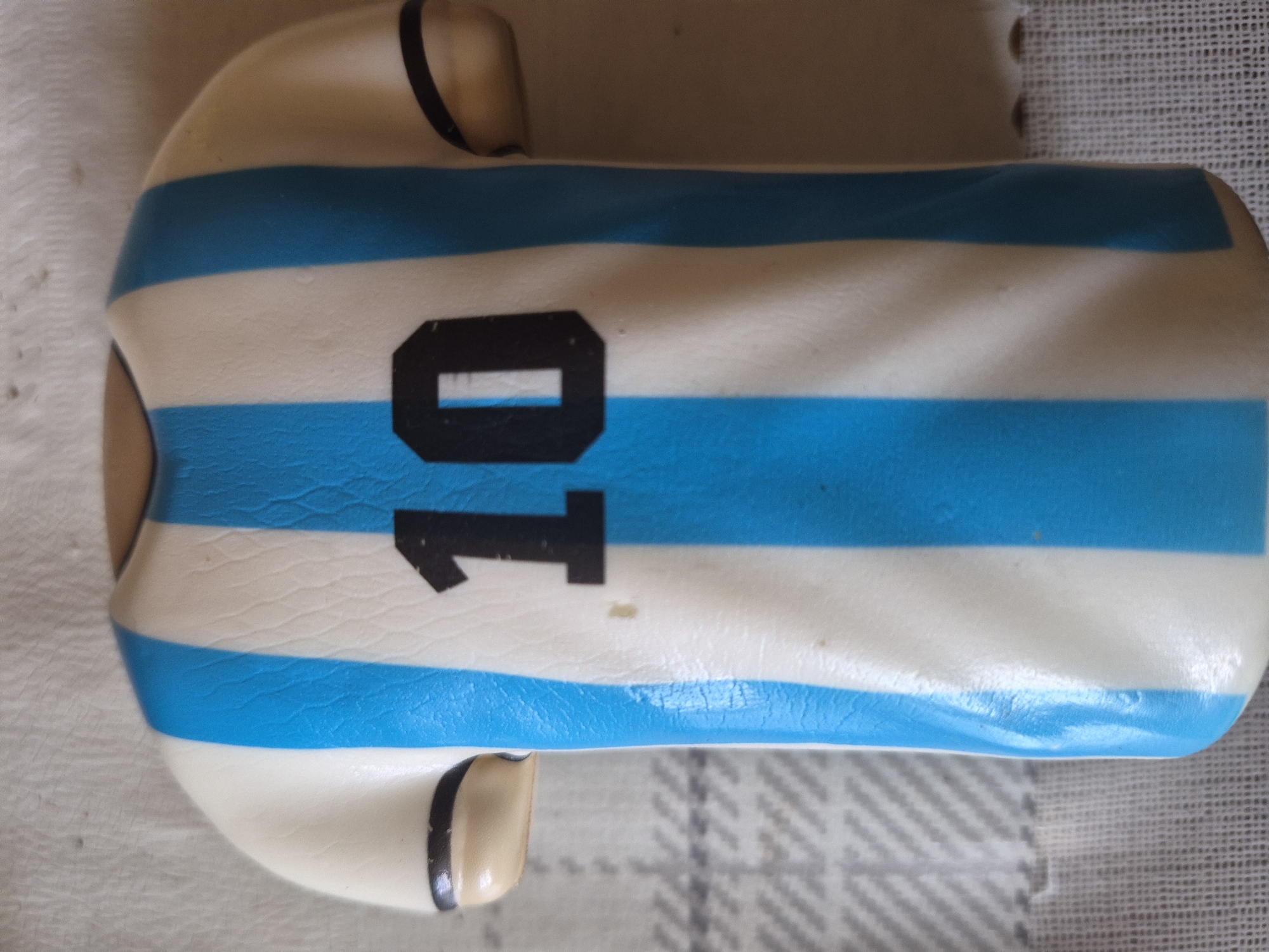 The image displays a close-up, top-down view of a miniature, stylized object resembling a football (soccer) jersey, oriented vertically with its top to the left of the frame. The object is predominantly white with three prominent light blue horizontal stripes across its body, characteristic of the Argentina national football team's kit. On the back of this jersey-like item, the number "10" is clearly visible in black, with the "0" featuring a rectangular cutout in its center. Small black trim is visible around the collar and short sleeves, which are molded as part of the overall form.
The object appears to be made of a smooth material, possibly plastic, ceramic, or a confectionery element like fondant, given its slightly glossy finish and molded contours. There are minor textural imperfections, particularly on the blue striped areas. The background consists of a light-colored, textured fabric, featuring both a ribbed pattern on the left and a subtle gray and white checkered design on the right. The lighting is soft and even, suggesting an indoor setting. While the location context is Yerevan, Armenia, no specific visual cues related to the city are discernible within the image itself. The item appears to be a decorative piece or collectible, possibly a cake topper, related to football fandom.