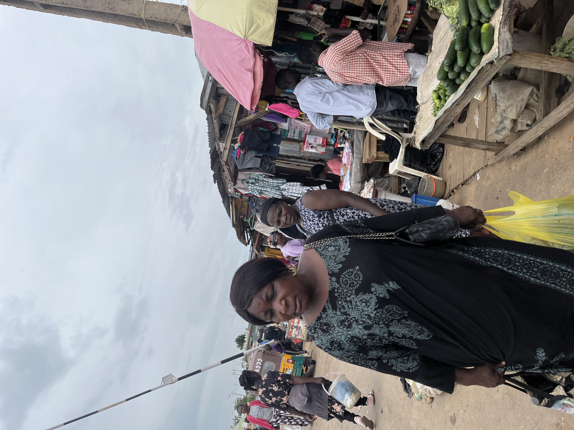 In the city of Gura Top, Nigeria, an outdoor market scene unfolds under a cloudy sky. A woman in a black, patterned dress walks forward, her gaze downcast. She carries a black purse on her left shoulder and a yellow plastic bag in her right hand. Behind her, another woman in a black and white patterned dress walks, her expression also unreadable.
Further back, several other individuals are visible. A man in a light blue striped shirt stands near a market stall filled with clothing. Another man in a red and white checkered shirt stands beside him, looking at his phone. A variety of goods are displayed at the stalls, including clothing and fresh produce like cucumbers. In the background, a small three-wheeled vehicle, commonly known as a Keke Napepa, is parked. The ground is dirt and uneven, typical of market areas. The overall atmosphere suggests a busy, everyday market day.