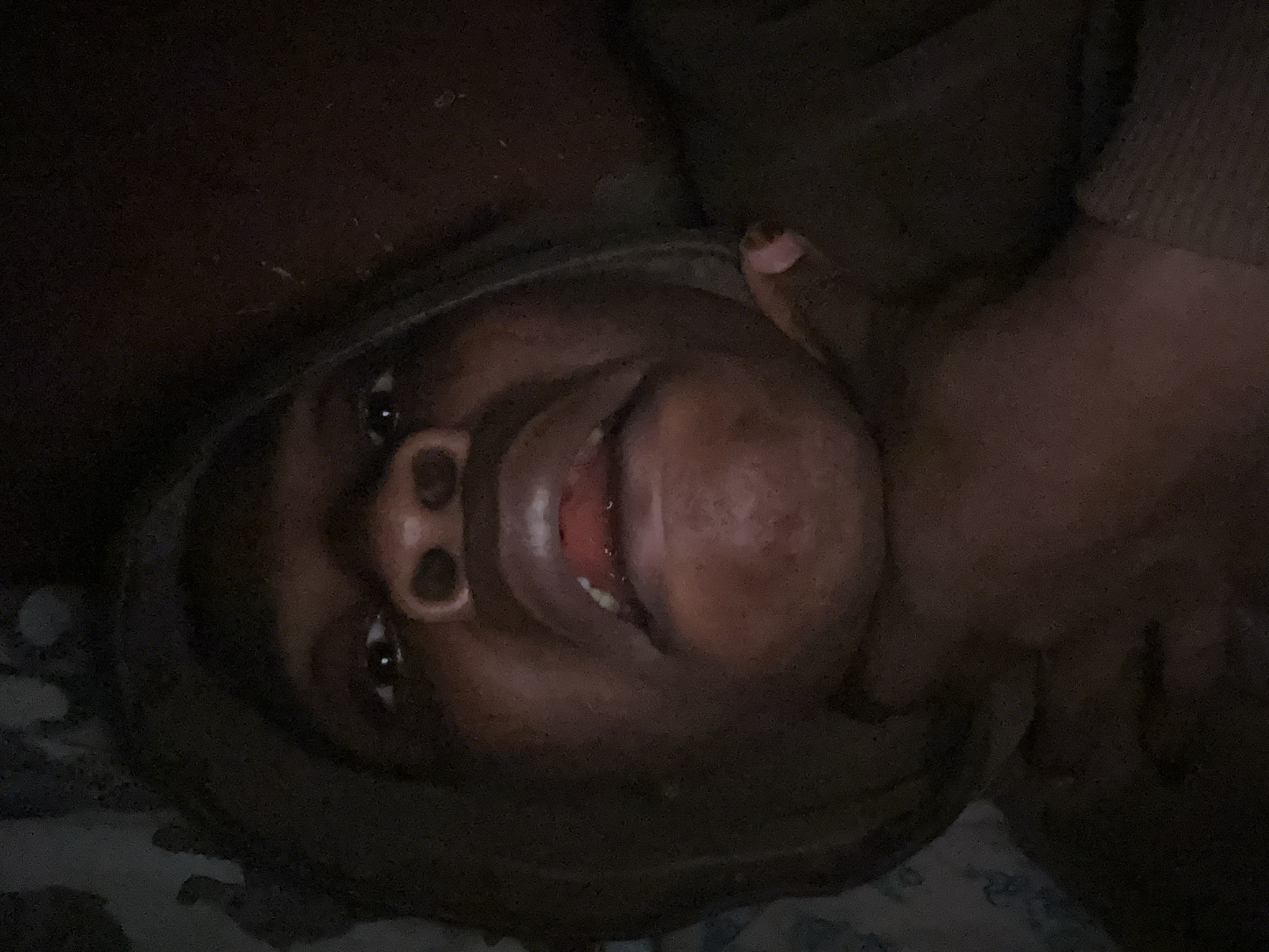 The image shows a close-up, dimly lit, overhead shot of a person wearing a hooded garment, appearing to be in a relaxed or reclining position. The person's face is the primary focus, with their mouth slightly open, revealing a glint of red on their lower lip. A hand is visible near the person's chin, with a pink fingernail. The background is dark and indistinct, with a patterned fabric visible to the left. The overall impression is of a candid, personal moment captured in low light. There is no identifiable information in the image that would suggest a specific location in Ikorodu, Nigeria.