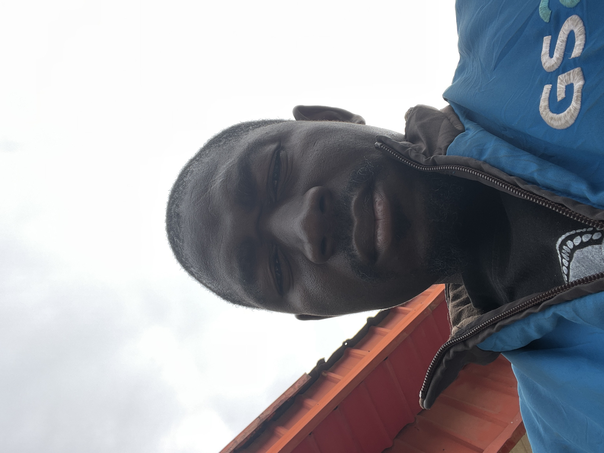 A low-angle, close-up, eye-level outdoor selfie captures a Black man from the chest up against an overcast sky. He is wearing a blue jacket with "GS" embroidered in white and teal on the right side of his chest, over a black t-shirt. A portion of an orange corrugated metal roof structure is visible on the left side of the frame. The image was taken in Jos, Nigeria.