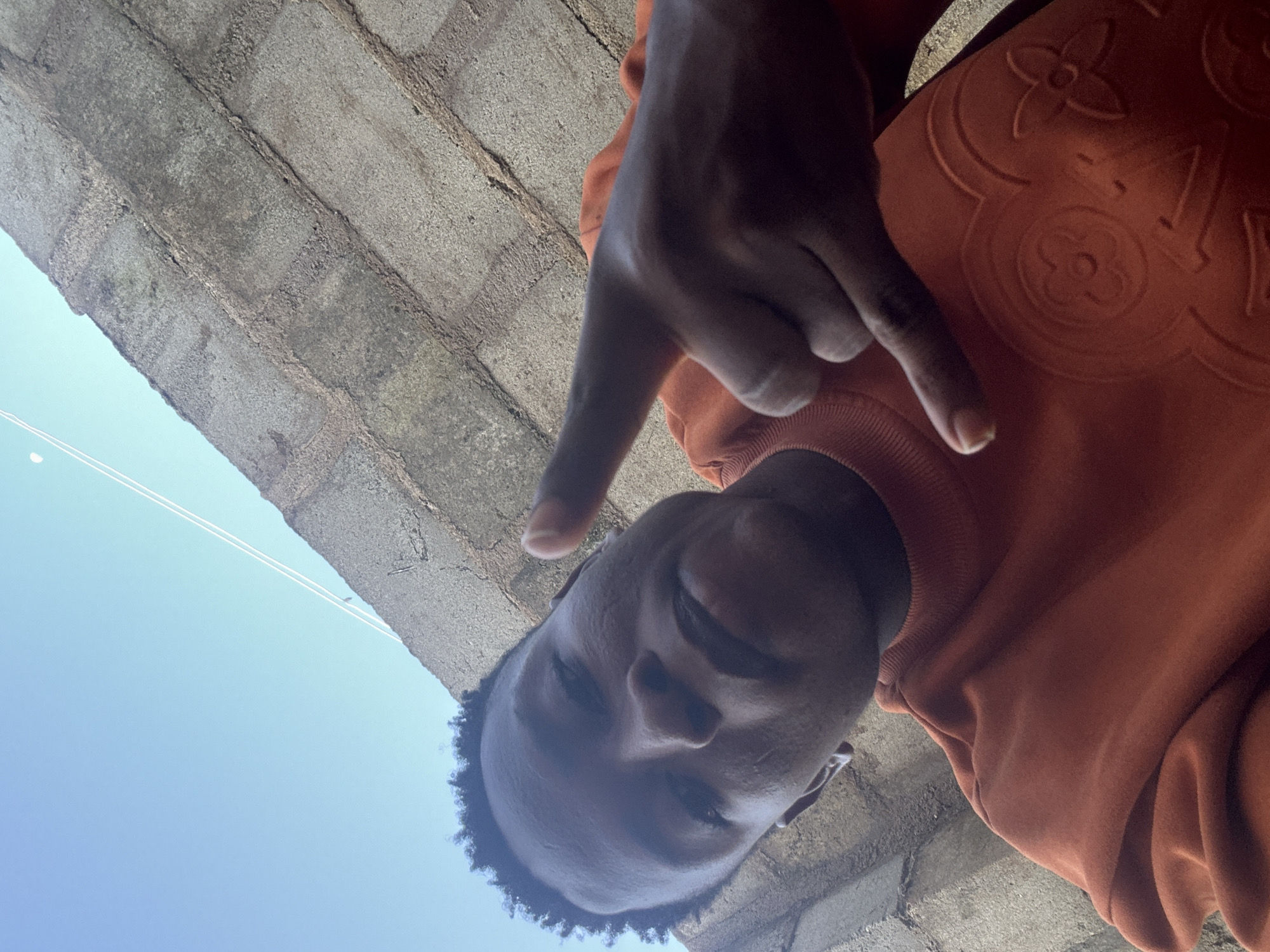 This is a low-angle, close-up shot of a young Black person posing for a selfie outdoors. The person is wearing an orange sweatshirt with a Louis Vuitton-inspired pattern embossed on it. They are making a "V" hand sign near their face. The background consists of a brick wall and a bright blue sky with thin white lines, possibly representing power lines or clouds. The lighting suggests it is daytime. The image has an upward-tilted perspective, making the viewer look up at the subject and the sky. The person's expression appears neutral or slightly amused. The scene is likely in a residential or outdoor area in Ikorodu, Nigeria, given the context. No discernible text is visible in the image.