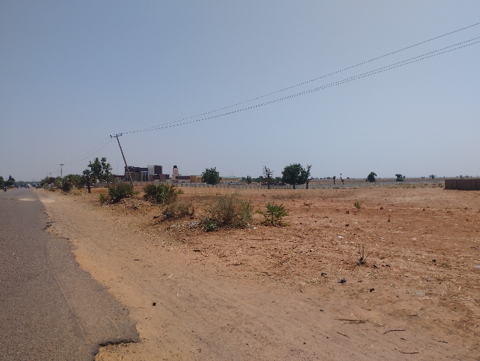 The scene is a dirt road on the outskirts of Sangaro, Nigeria, with sparse vegetation. The road is paved in the foreground and extends to the horizon. There is a small amount of traffic on the road. The road is bordered by a ditch. The area is dry and dusty. There are trees in the background, and the sky is clear. There are power lines running across the image.