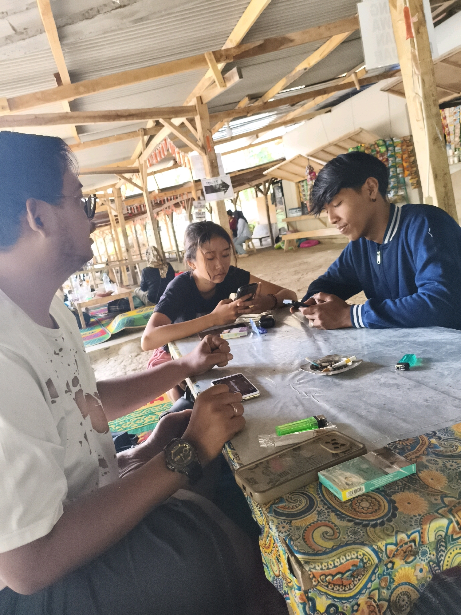 Three individuals are seated at a table covered with a patterned tablecloth, likely in an outdoor or semi-outdoor establishment in Babakan Madang, Indonesia. The table is made of a light-colored material, possibly plastic or laminate, and is partially covered by a clear plastic sheet.
On the left, a person wearing a white t-shirt with distressed detailing and sunglasses is visible from the chest up, looking to their left. They are wearing a wristwatch with a dark strap and a circular face.
In the center, a young woman with dark hair pulled back is looking down at her phone. She is wearing a black t-shirt and appears to be seated on the floor or a low seating arrangement.
To the right, a young man in a blue jacket with white stripes is looking down at his phone. He has dark hair and is wearing an earring.
On the table, various objects are present, including mobile phones, a cigarette pack, an ashtray with cigarette butts, a green lighter, and what appears to be a cigarette packet. The background suggests a casual setting with wooden beams and a corrugated metal roof, consistent with a roadside stall or a simple eatery in a tropical environment. There are also signs in the background, one of which appears to indicate "TOILET". The ground beneath the table is sandy or dirt.