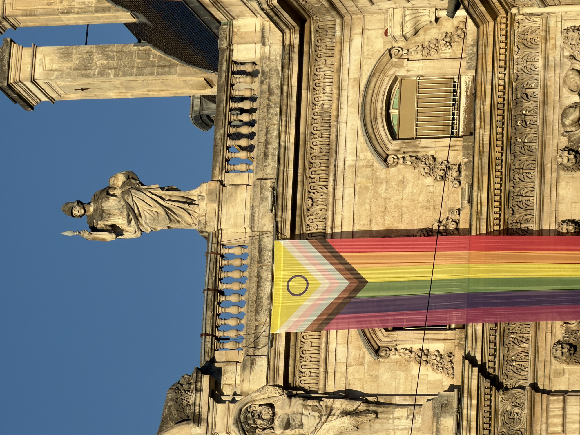 A large, colorful banner, resembling a progress pride flag, is draped over the side of a stone building, a clear blue sky serving as the backdrop. The building features classical architectural elements, including ornate carvings and statues, and it appears to be a historical structure. One statue, at the top of the building, depicts a robed figure holding a spear and a sphere. Below it, a balustrade runs horizontally, and a prominent banner hangs down, displaying stripes of various colors including yellow, white, pink, purple, green, and red, with a purple circle emblem at the top. The banner is suspended by a metal rod. Further down, carved lion heads are visible on the facade. The image appears to have been taken during the day, with sunlight illuminating the stone structure. The specific location is the Hôtel de Ville (City Hall) in Lyon, France.