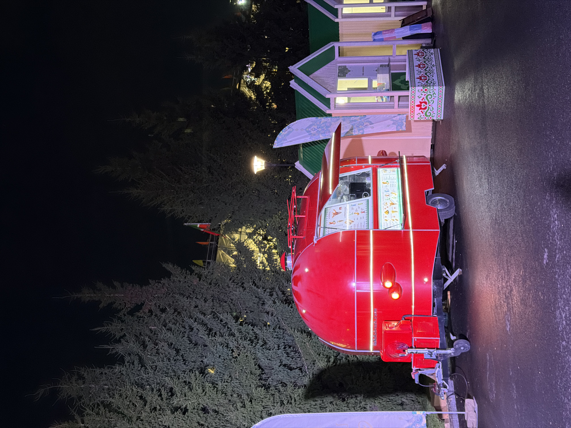A red food truck with a rounded, retro design is parked on a dark, wet asphalt surface. The truck is illuminated by white LED lights that outline its shape. A menu board with illustrations of food items is visible on the side of the truck, along with a small awning extending from the serving window. In the background, several small, gabled structures with green roofs are lined up, appearing to be market stalls or kiosks. A tall street lamp with a warm yellow light stands to the left of the food truck. Behind the stalls, evergreen trees are visible against a dark night sky, with some lights of a building visible through the trees. Several flags are also flying in the distance. The scene suggests an outdoor event or market taking place at night.