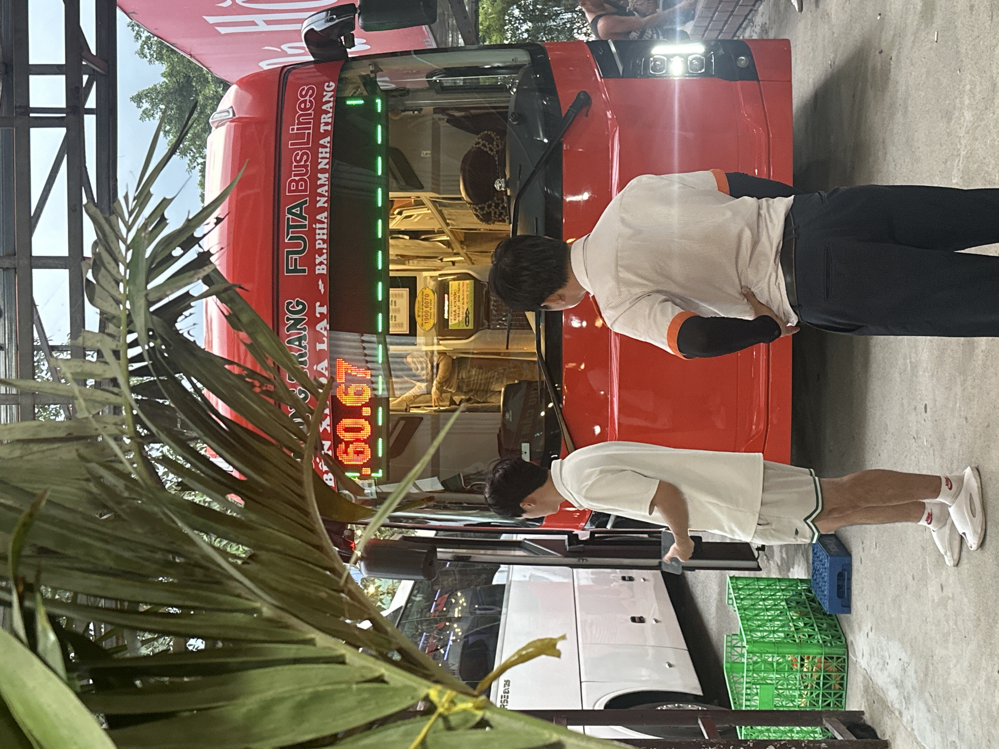 A red FUTA Bus Lines bus, with the route "BX. PHIA NAM NHA TRANG," is parked. The number "1900 6070" is displayed inside the bus. Two men stand in front of the bus, with a palm frond partially obscuring the view. One man wears a white shirt with orange accents and dark pants, while the other wears a white t-shirt and light-colored shorts. Crates, some green and some blue, are stacked nearby. A white van is partially visible behind the bus. The digital display above the windshield appears to read "60.67". The location is in Vietnam, in a place written "BX. PHIA NAM NHA TRANG".